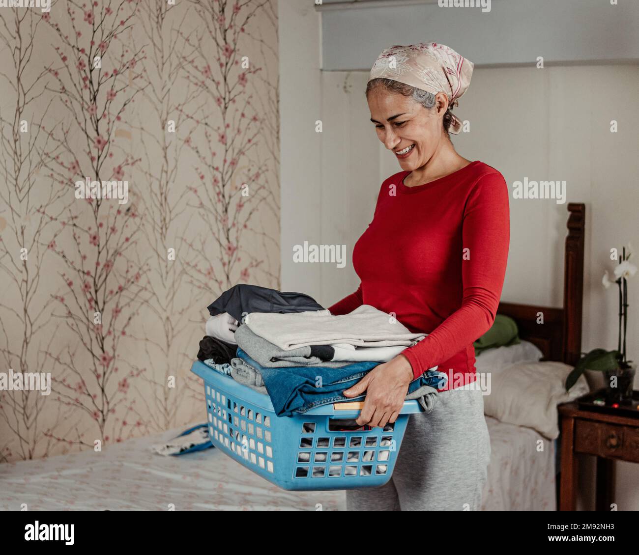 Positiv reife Frau in lässigem Outfit lächelt und trägt einen blauen Korb mit Stoff nach der Wäscherei zu Hause Stockfoto