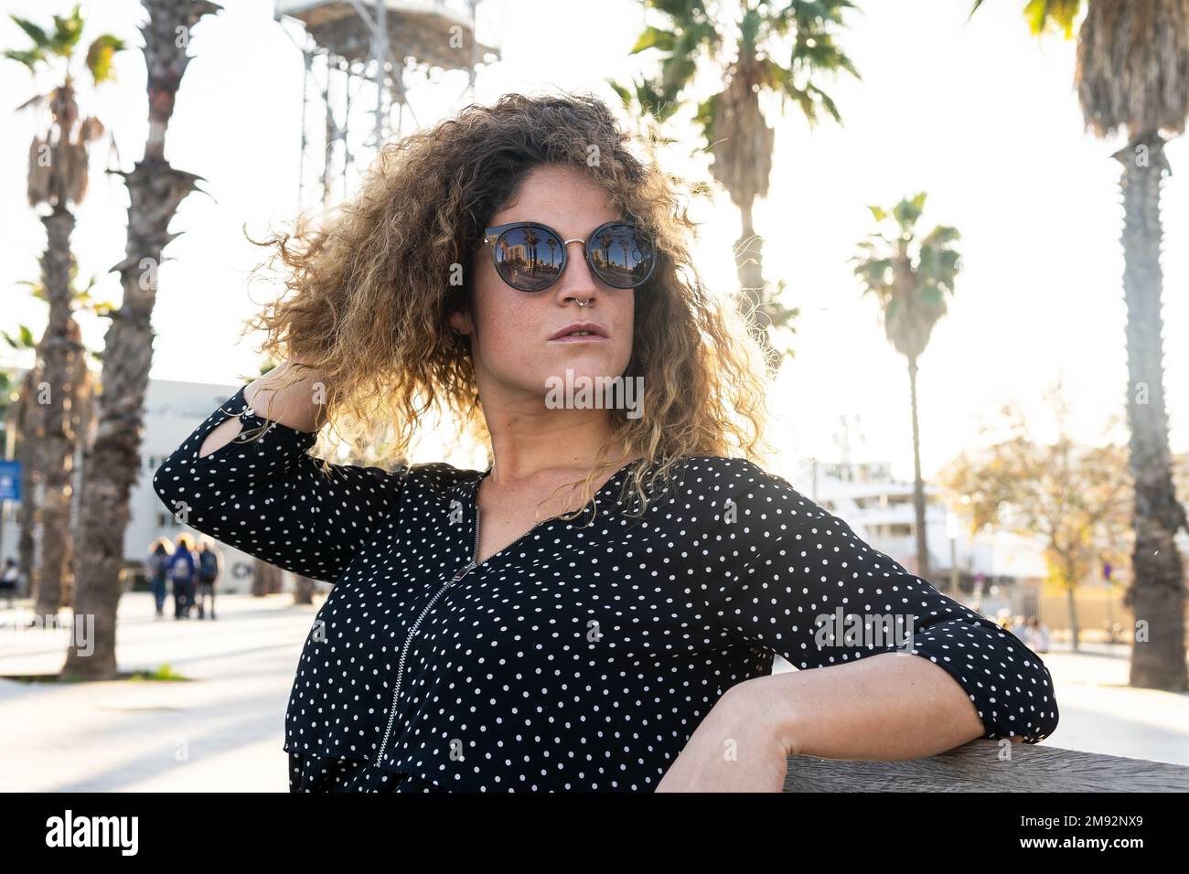 Frau mit Sonnenbrille und schwarzem gepunktetem Hemd, die an sonnigen Tagen am Wochenende auf einer Bank in der Nähe hoher Palmen ruht Stockfoto