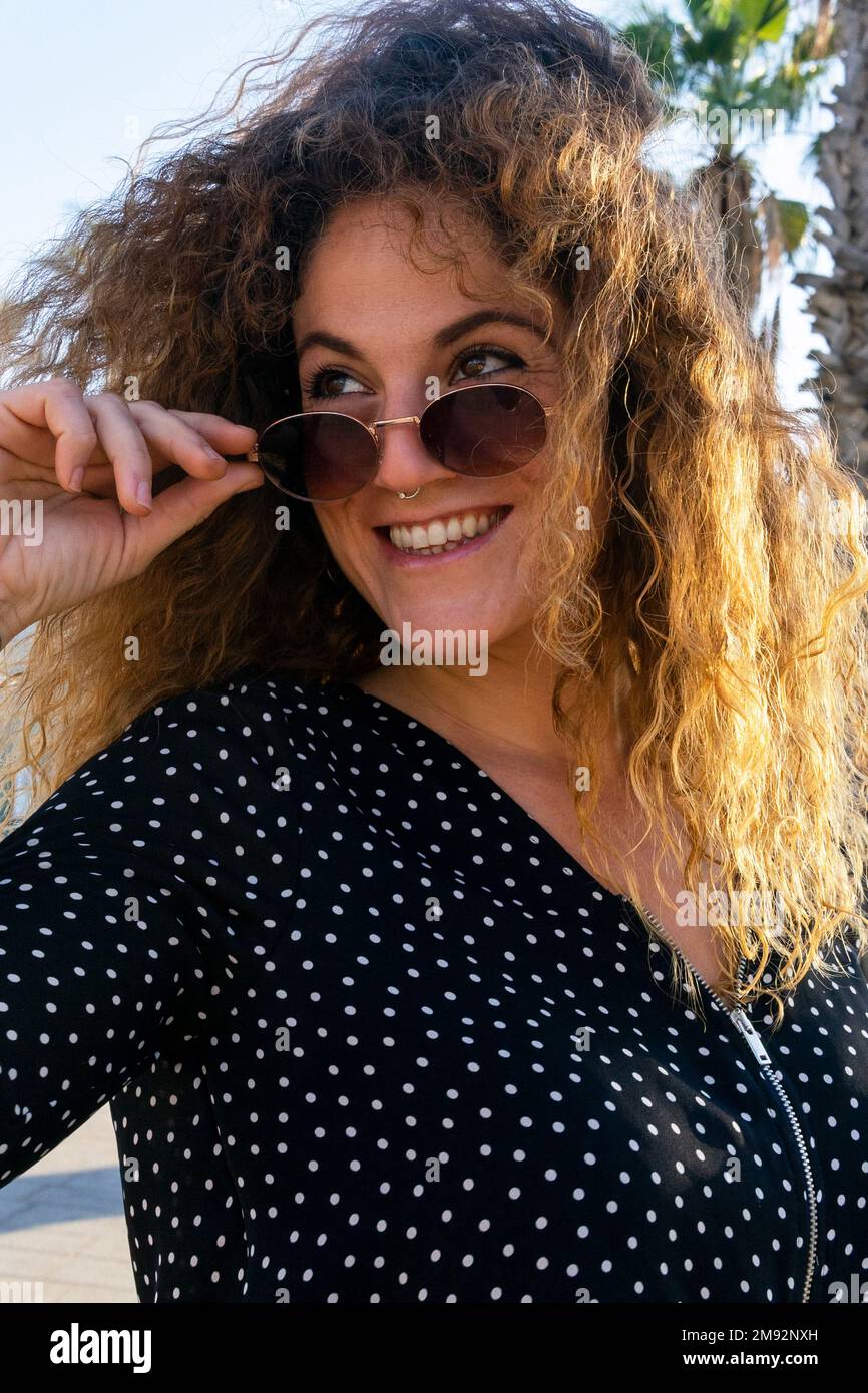 Fröhliche junge Frau mit lockigem Haar in lässiger Kleidung, die lächelt und wegschaut, während sie die Sonnenbrille in der Nähe von Handflächen justiert Stockfoto