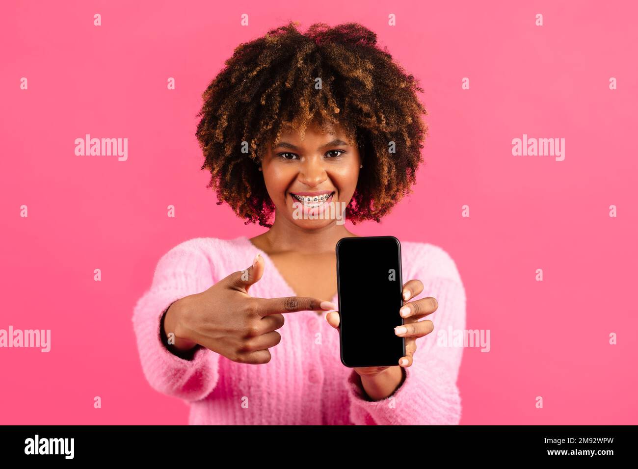 Die fröhliche, afroamerikanische Dame mit Zahnspange zeigt den Finger am Telefon mit leerem Bildschirm Stockfoto