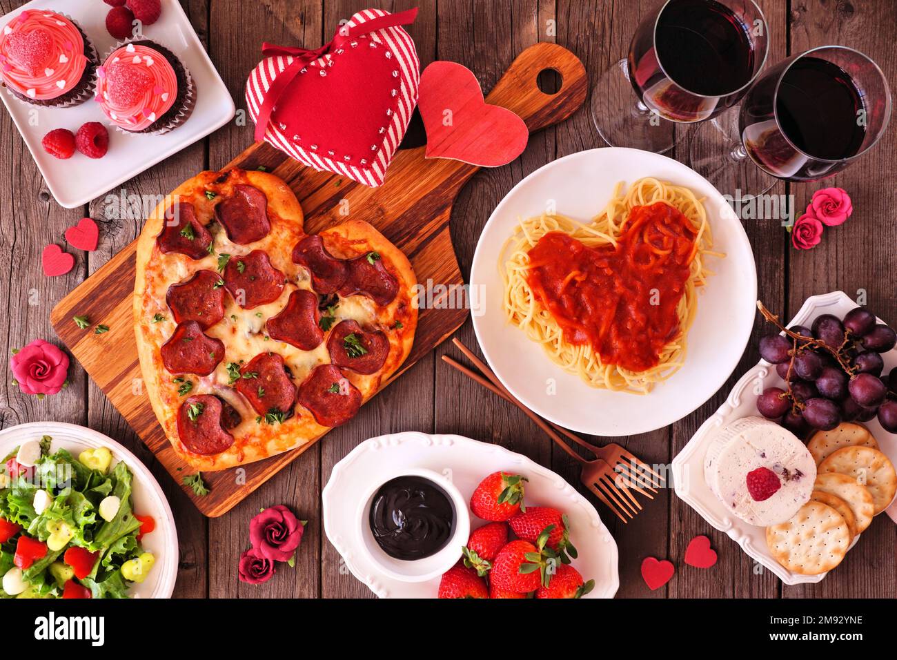 Hausgemachtes Abendessen am Valentinstag. Tischeinstellung von oben auf dunklem Holzhintergrund. Herzförmige Pizza, Pasta, Wein, Käseplatte und Desserts. Stockfoto