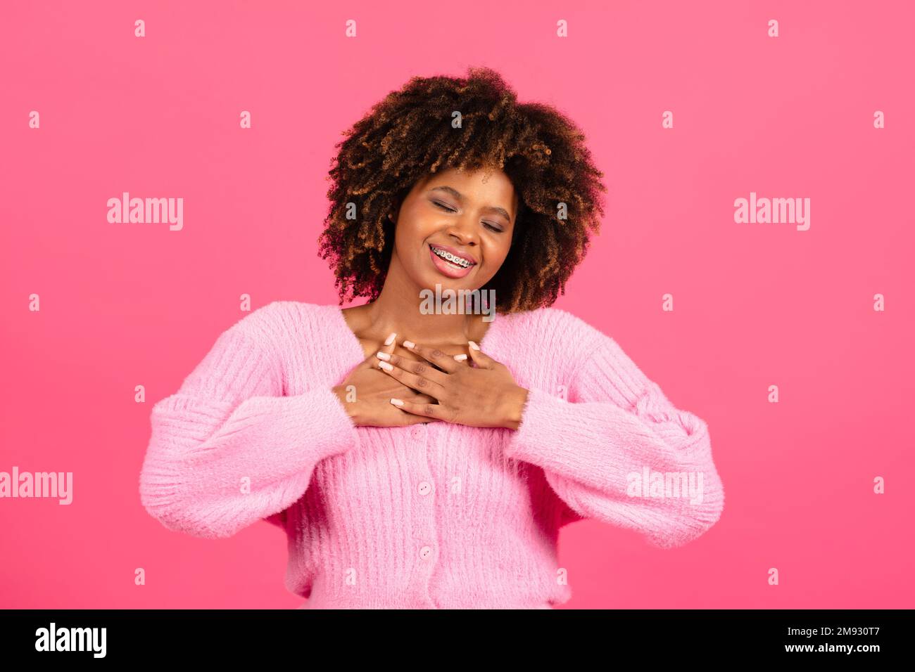 Lächelnde junge, afroamerikanische, lockige Frau in legerer Kleidung drückt die Hände auf die Brust, genießen Sie es dankbar Stockfoto