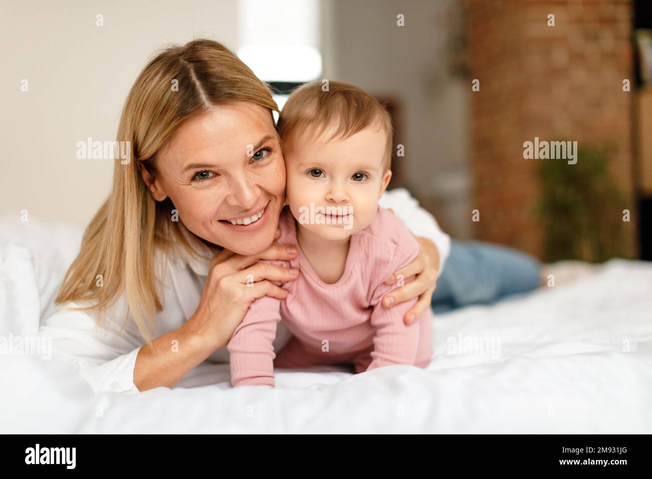 Glückliche Mutterschaft. Mami spielt mit einem niedlichen Kleinkind, das auf dem Bett krabbelt, vor der Kamera lächelt, das Schlafzimmer ist innen Stockfoto