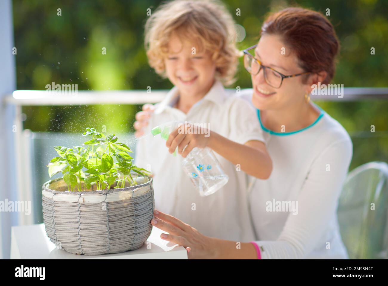 Frau und Kind, Wasser, Balkonpflanze. Kräutergarten auf der Terrasse. Junge Mutter und Kind sprühen Wasser auf Topfkräuter. Basil in einem Topf auf der Apartmentterrasse. Stockfoto