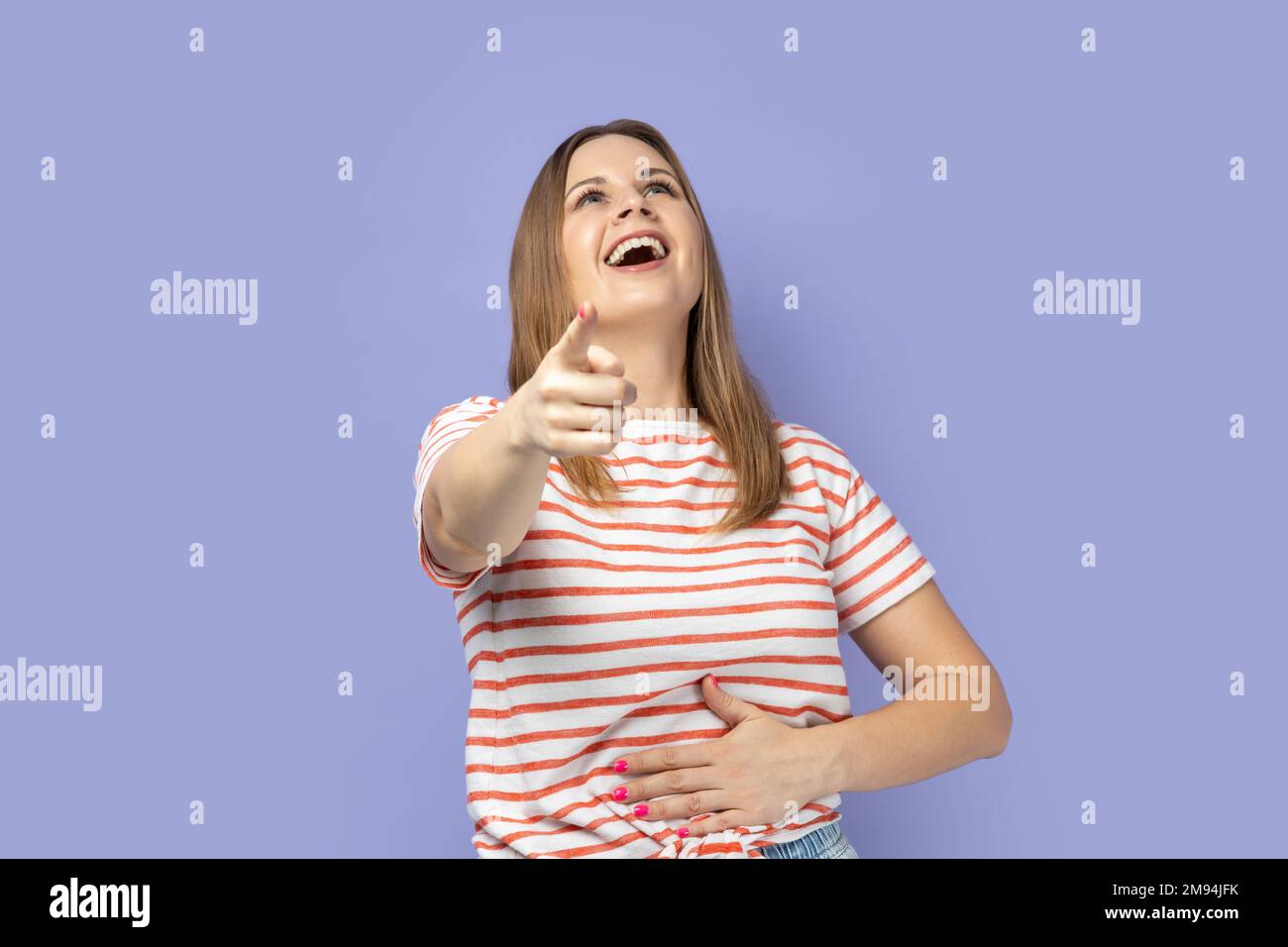 Ha ha, du bist lächerlich. Porträt einer Frau, die ein gestreiftes T-Shirt trägt und mit dem Finger auf die Kamera zeigt, laut lacht und lustige Freundinnen aufzieht. Studio-Aufnahme im Innenbereich isoliert auf lila Hintergrund. Stockfoto