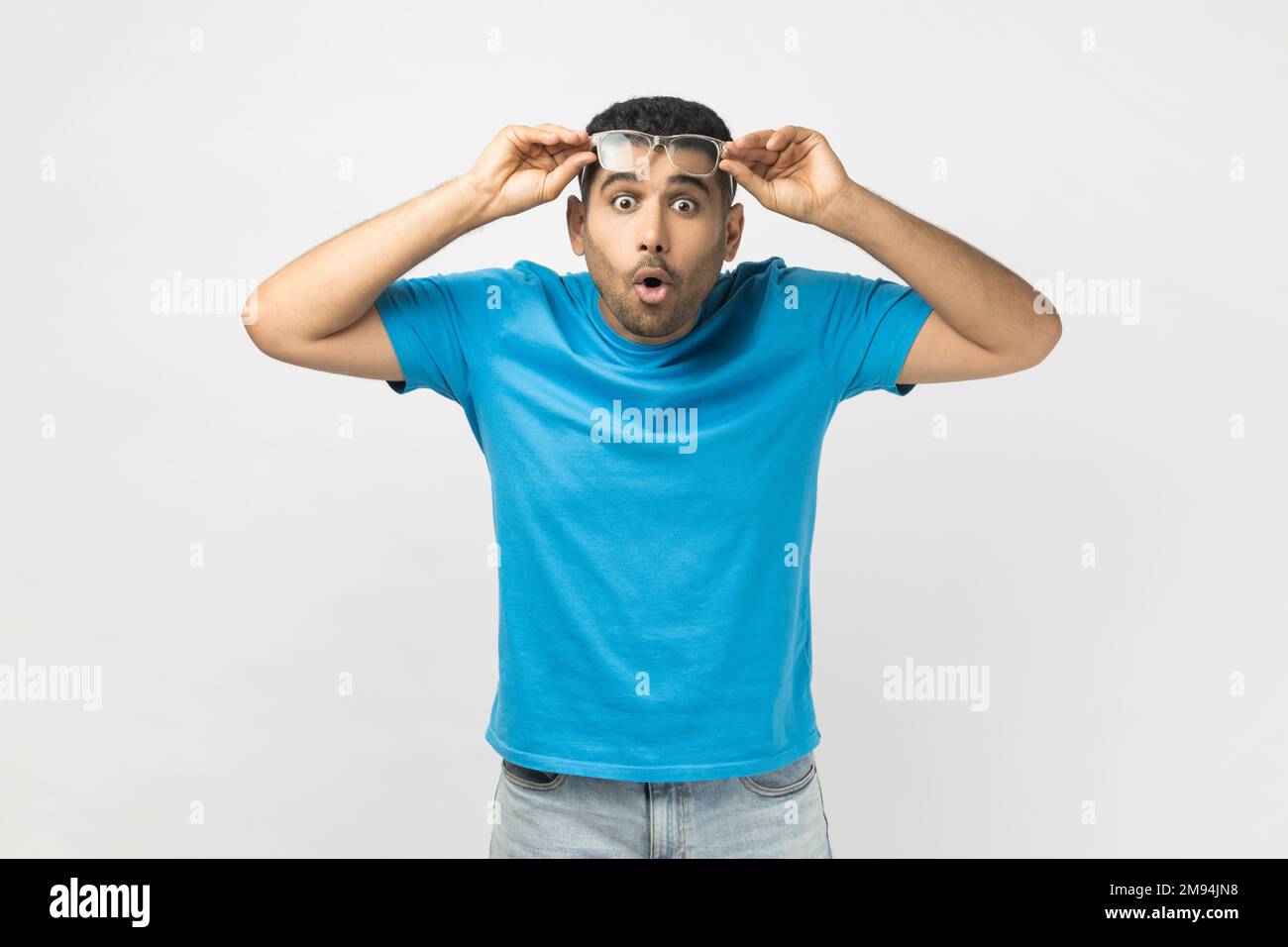 Portrait eines schockierten, erstaunten, unrasierten Mannes, der ein blaues T-Shirt trägt, seine optische Brille hochhob, sieht etwas Überraschtes. Studioaufnahmen im Innenbereich isoliert auf grauem Hintergrund. Stockfoto