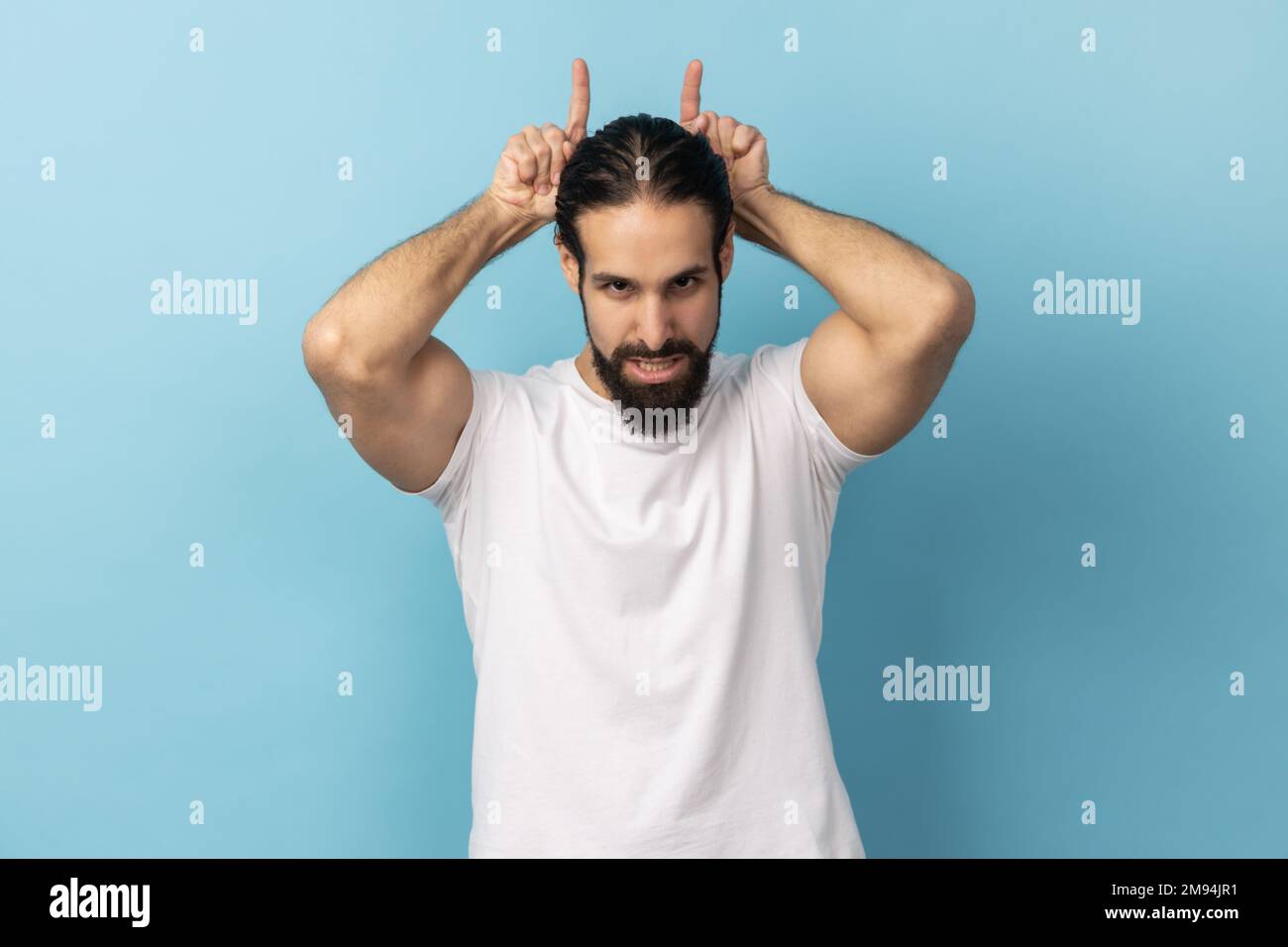 Porträt eines aggressiven Tyrannen mit Bart und weißem T-Shirt, mit Stierkopfgesten über dem Kopf, Stirnrunzeln wie vor dem Angriff, Indoor Studio-Aufnahme isoliert auf blauem Hintergrund. Stockfoto