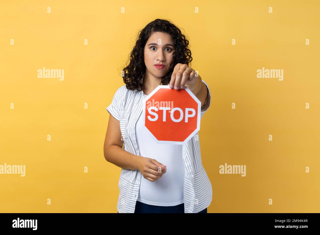 Porträt einer ernsten Frau mit dunklem, welligem Haar mit rotem Stoppschild, das auf die Kamera schaut, mit ernstem Gesichtsausdruck, Verbot. Innenstudio-Aufnahme isoliert auf gelbem Hintergrund. Stockfoto