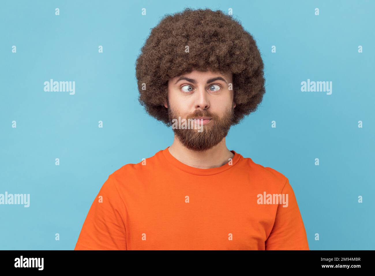 Portrait eines verrückten, lustigen Mannes mit Afro-Haarschnitt, der ein orangefarbenes T-Shirt trägt, mit gekreuzten Augen steht und mit einem albernen komischen Gesicht aussieht. Innenstudio, isoliert auf blauem Hintergrund. Stockfoto
