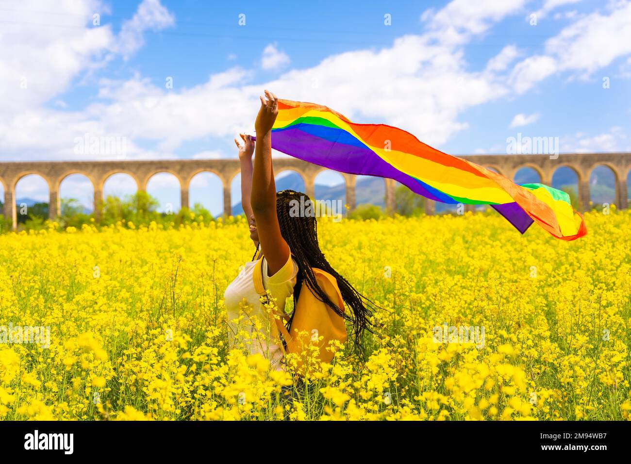 Ein schwarzes, ethnisches Mädchen mit Zöpfen, das die LGBT-Flagge in einem Feld gelber Blumen hält, Tag des Stolzes, Konzept der freien Frauen Stockfoto