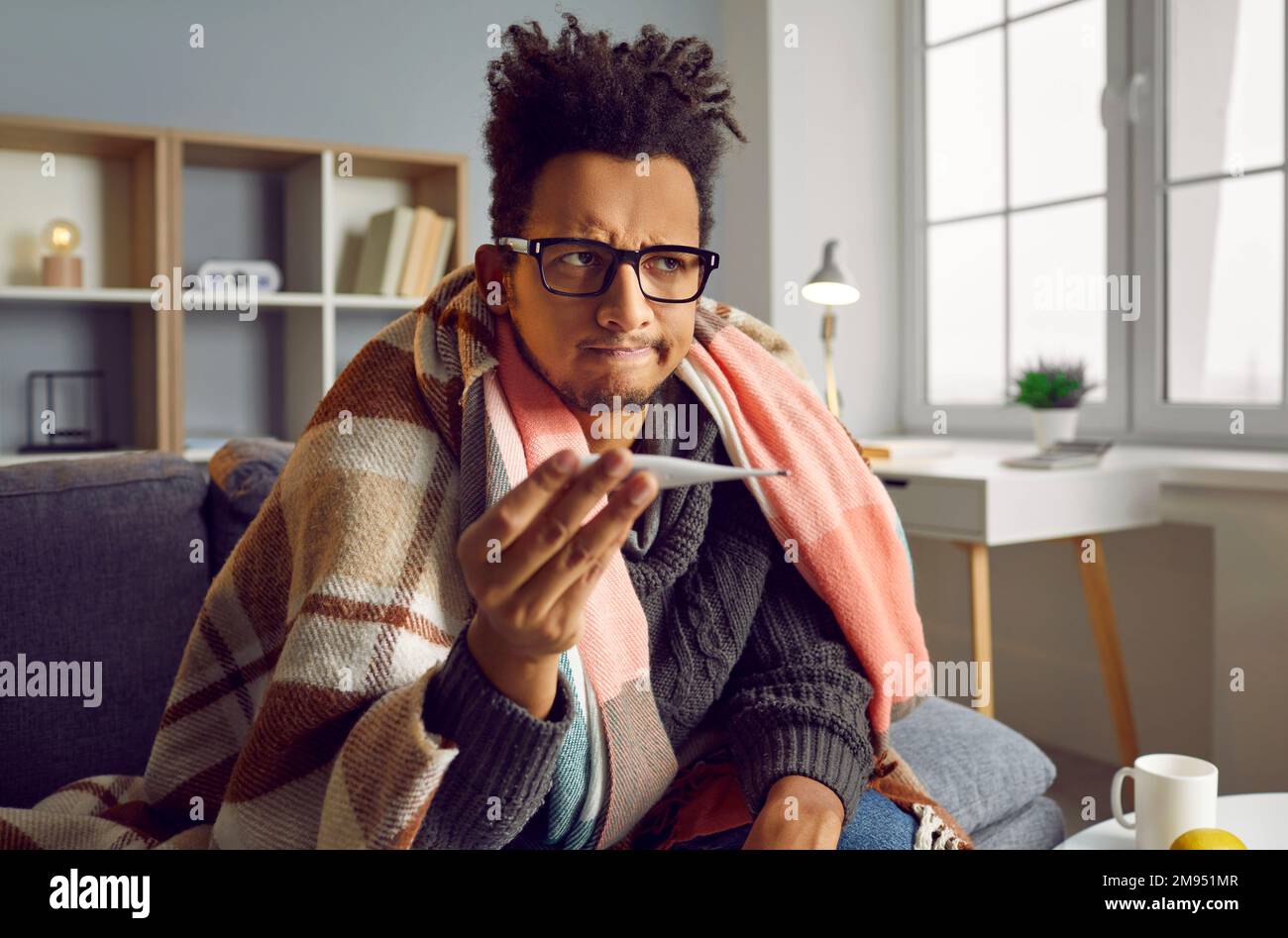 Kranker junger afroamerikanischer Mann, der seine Temperatur prüft, in einer Decke zu Hause verpackt. Stockfoto