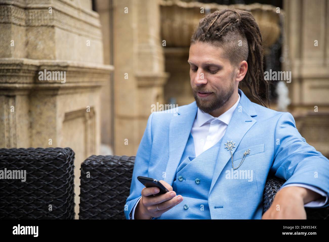 Ein junger erfolgreicher Mann im blauen Anzug, der in einem luxuriösen Straßencafé auf ein Smartphone schaut Stockfoto