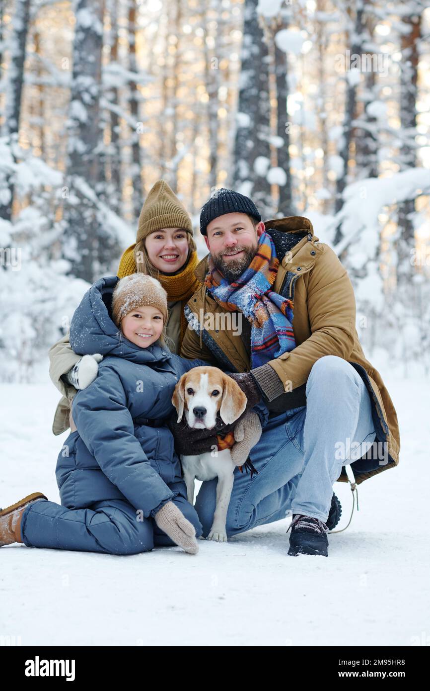 Porträt einer glücklichen Familie, die vor der Kamera lächelt, während sie mit dem Hund im Winterwald spazieren geht Stockfoto