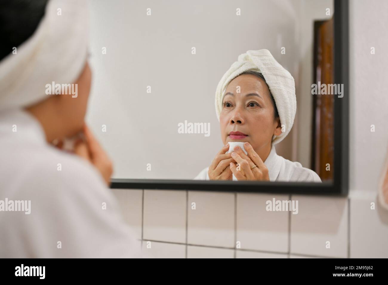 Wunderschöne 60s-jährige Asiatin, die ihr Make-up mit Make-up-Entferner und Wattepad entfernt, in den Spiegel schaut, sich auf die Dusche vorbereitet. Stockfoto