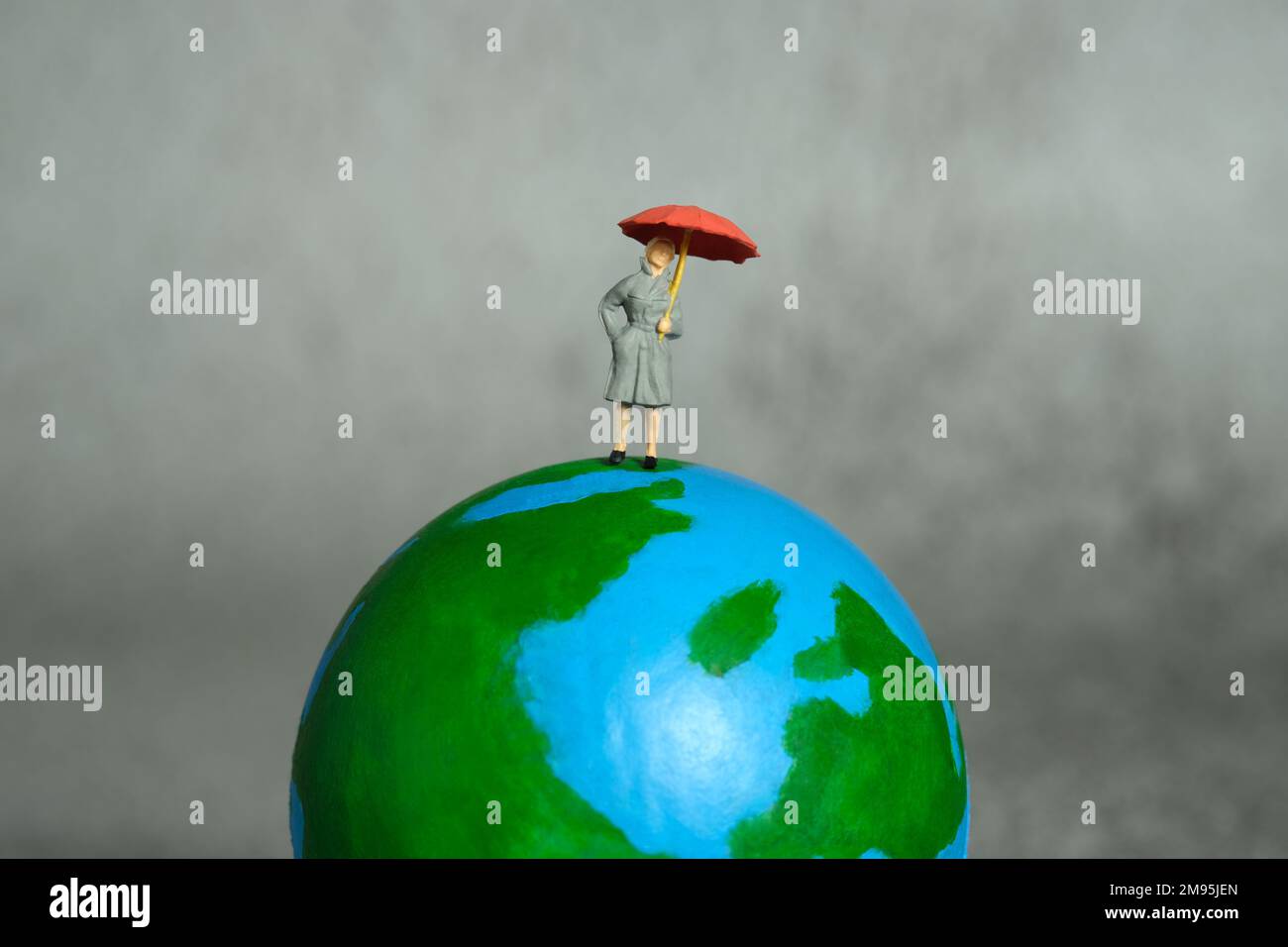 Miniatur-Menschen-Spielzeug-Figuren-Fotografie. Weibliche Nachrichtensprecherin, die über der Erde steht und die Wettervorhersage präsentiert. Grauer bewölkter Hintergrund. Bild-ph Stockfoto