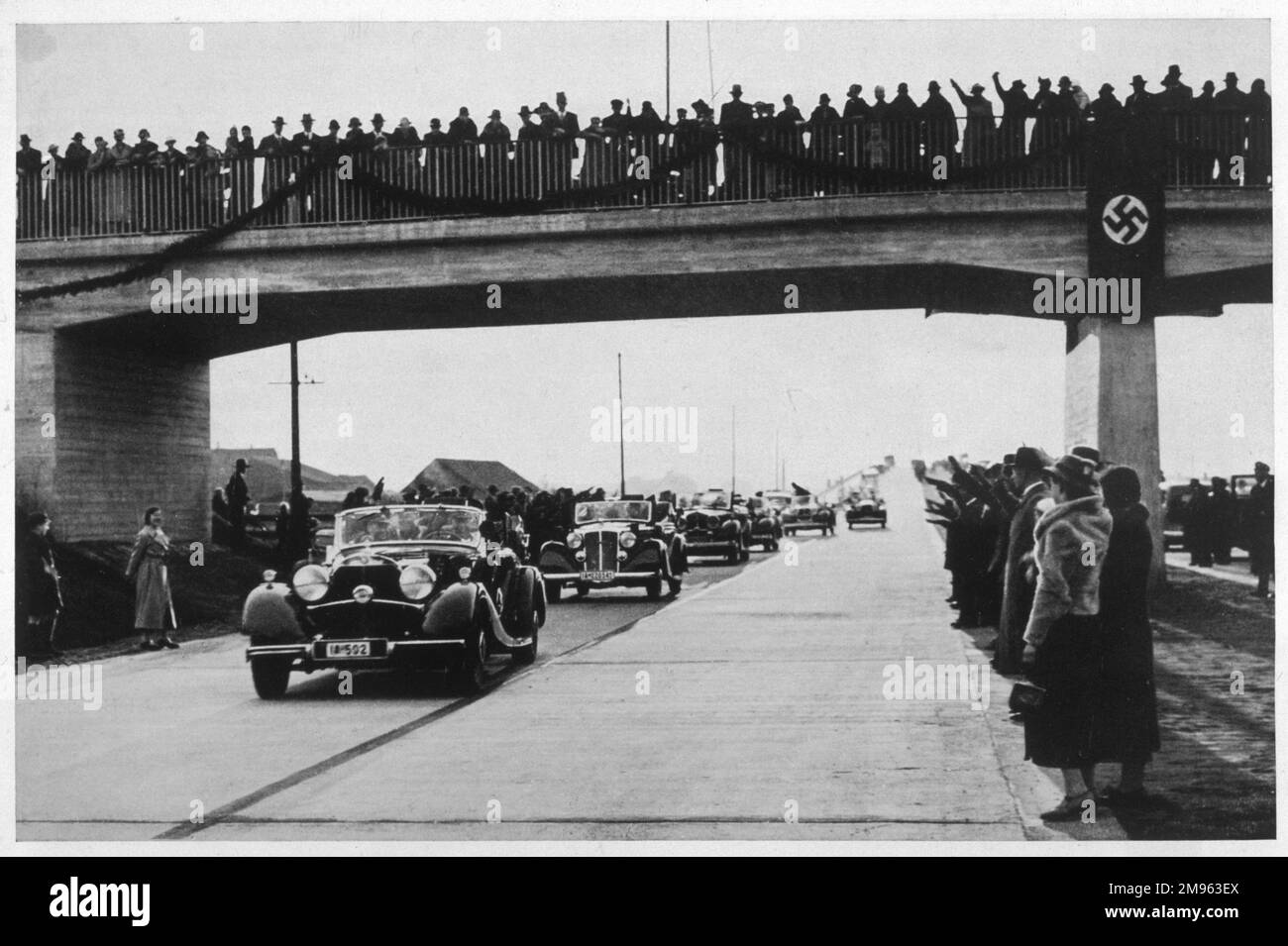 Aufgeregte Menschenmassen versammeln sich auf einer Brücke, um einer Reihe offizieller Nazi-Fahrzeuge auf einer neu eröffneten Autobahn zuzusehen. Stockfoto