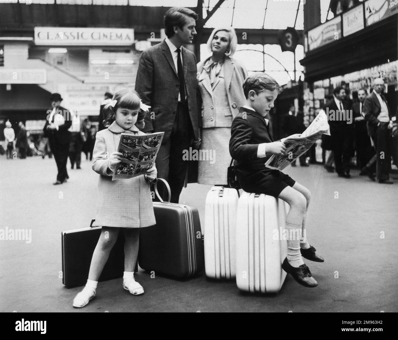 Eine Familie wartet am Bahnhof. Die Kinder sitzen auf ihren Koffern und lesen Comics, während sie warten. Stockfoto