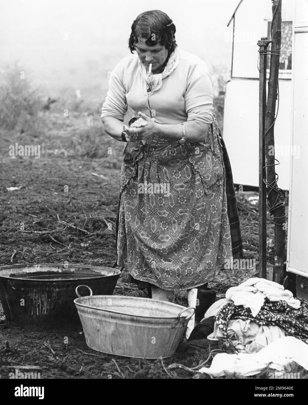Eine alte Zigeunerin trägt eine Schürze und raucht eine Zigarette, frisst sich die Hände, während sie sich darauf vorbereitet, einen Haufen Kleider von Hand in einem Metallbecken zu waschen. Stockfoto