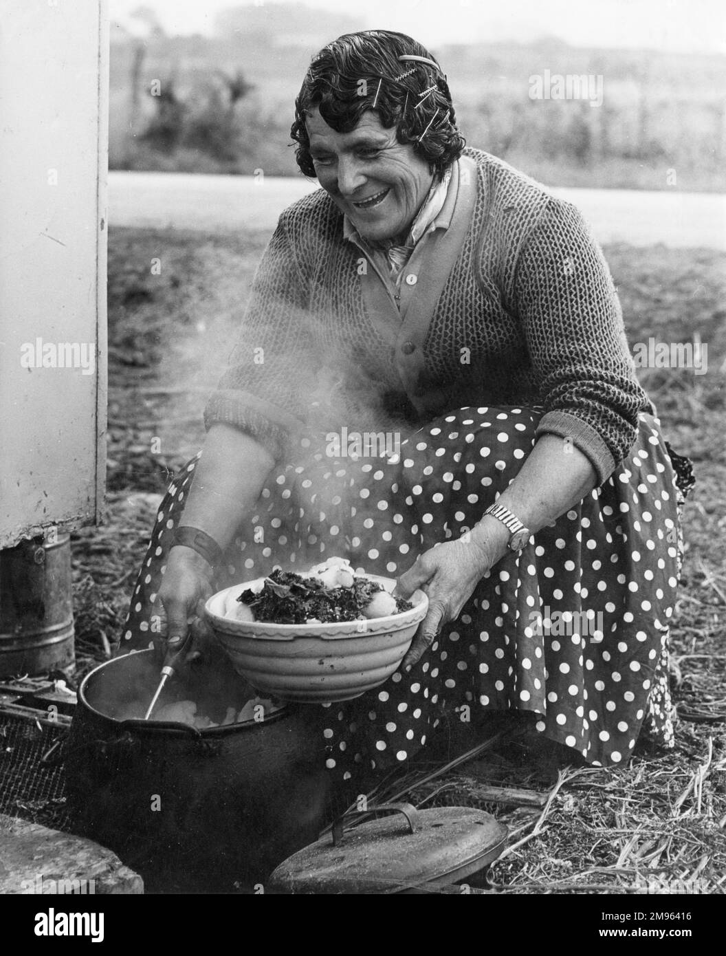Eine alte Zigeunerin lächelt, während sie Kartoffeln und Kohl aus einem kochenden Kessel serviert. Stockfoto