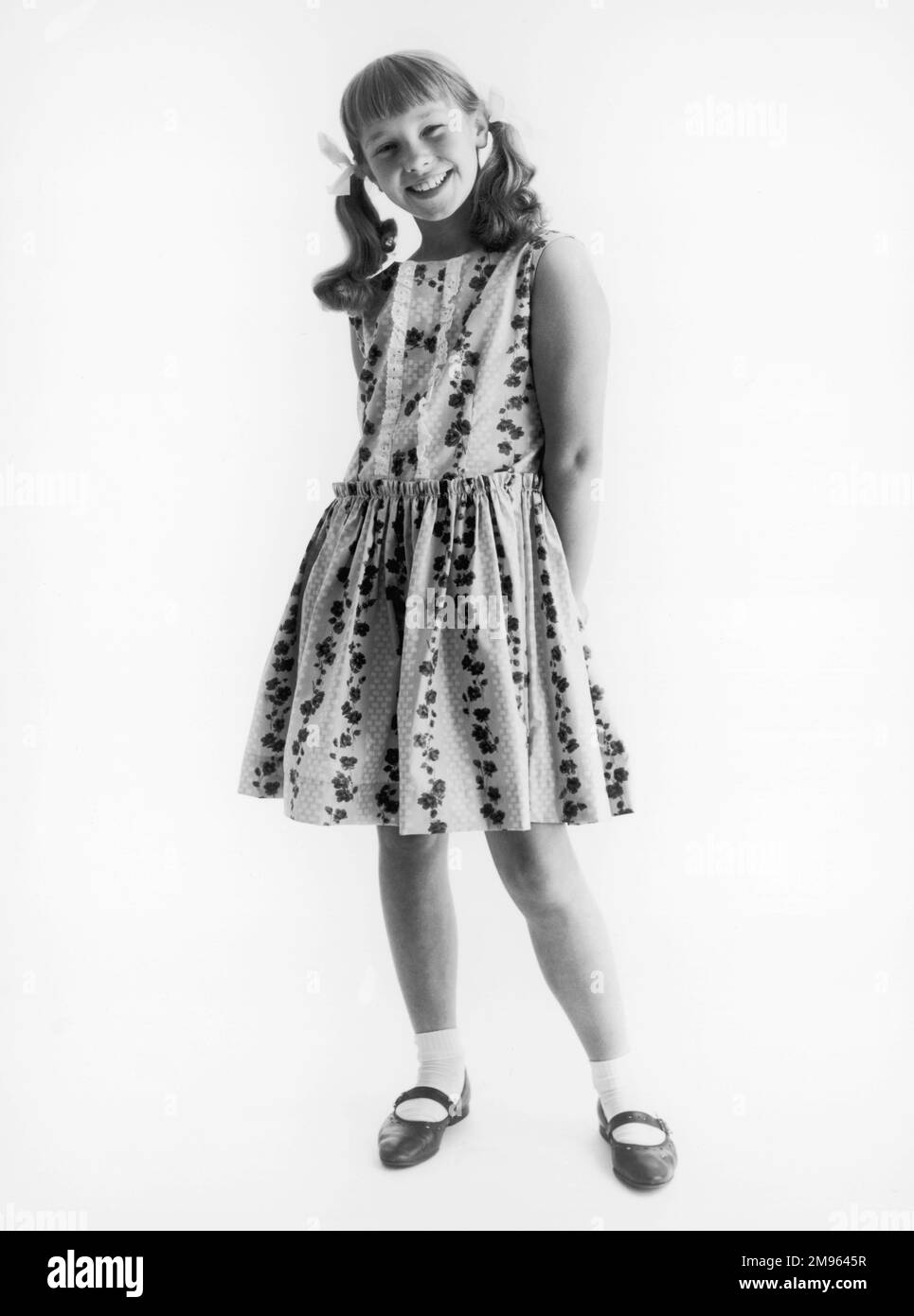 Ein Schulmädchen modelliert ein ärmelloses, geblümtes Sommerkleid, getragen mit Knöchelsocken und Sommerschuhen, ihr Haar in Zöpfen und Bändern. Stockfoto