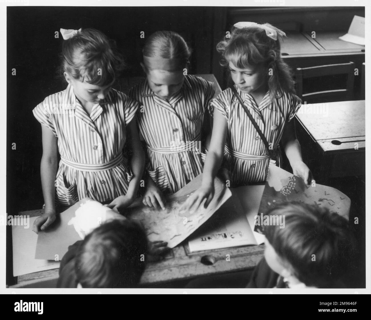 Drei Schulmädchen in Sommerkleidern schauen sich die Zeichnungen auf dem Schreibtisch vor ihnen genau an Stockfoto