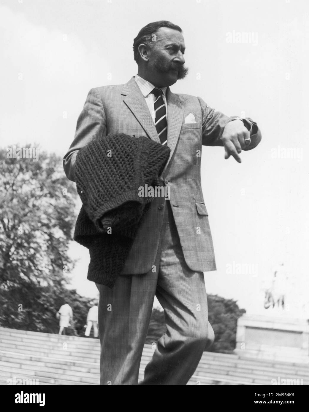 Ein mürrischer Gentleman, der auf seine Uhr schaut, um zu sehen, wie lange er schon spazieren geht. Es ist ein herrlicher Tag - er musste sogar seinen Wollspringer abnehmen. Stockfoto