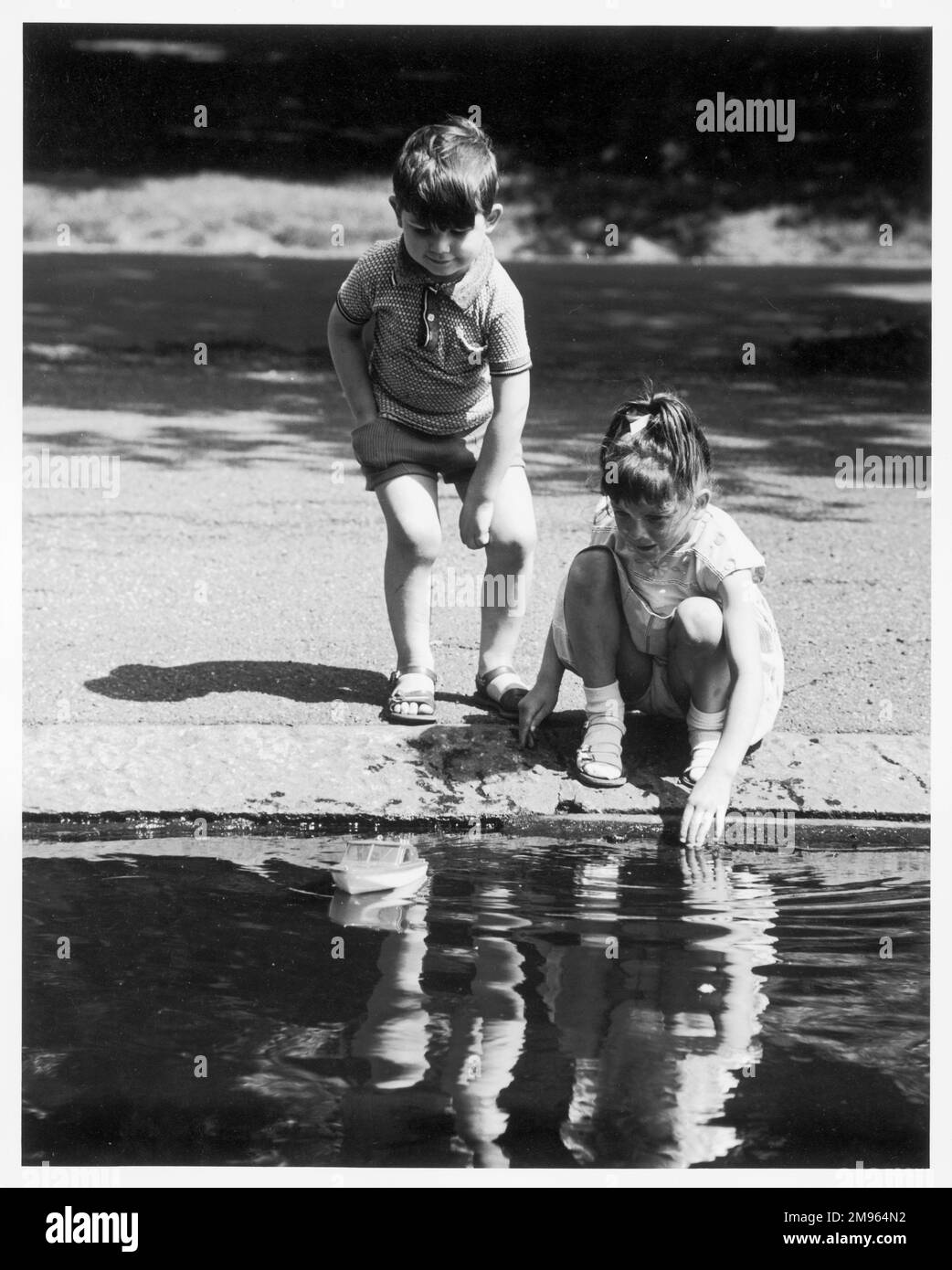 Zwei Kinder spielen mit einem Spielzeugboot auf einem Parksee. Stockfoto