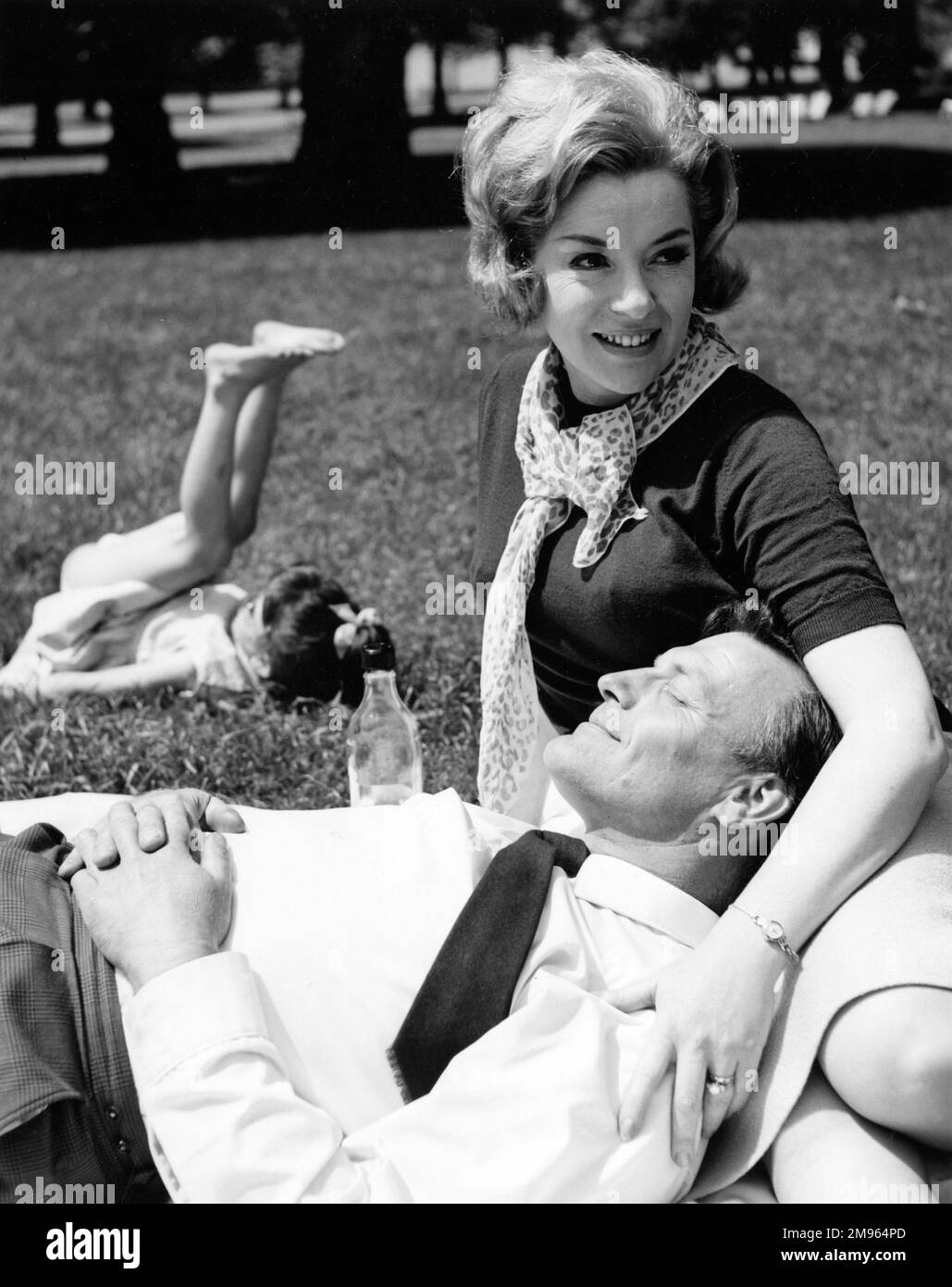 Ein Mann legt seinen Kopf auf den Schoß seiner Frau, während beide einen sonnigen Tag im Park mit den Kindern genießen, die im Hintergrund spielen. Stockfoto