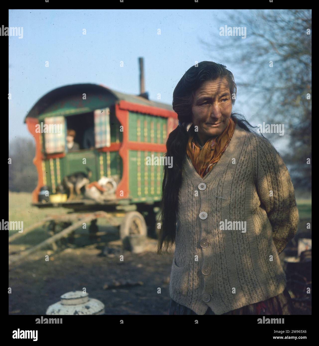 Eine ältere Zigeunerin mit langen dunklen Haaren und dicker Strickjacke steht vor der bunt bemalten Familienkarawane in einem Lager in Surrey Stockfoto