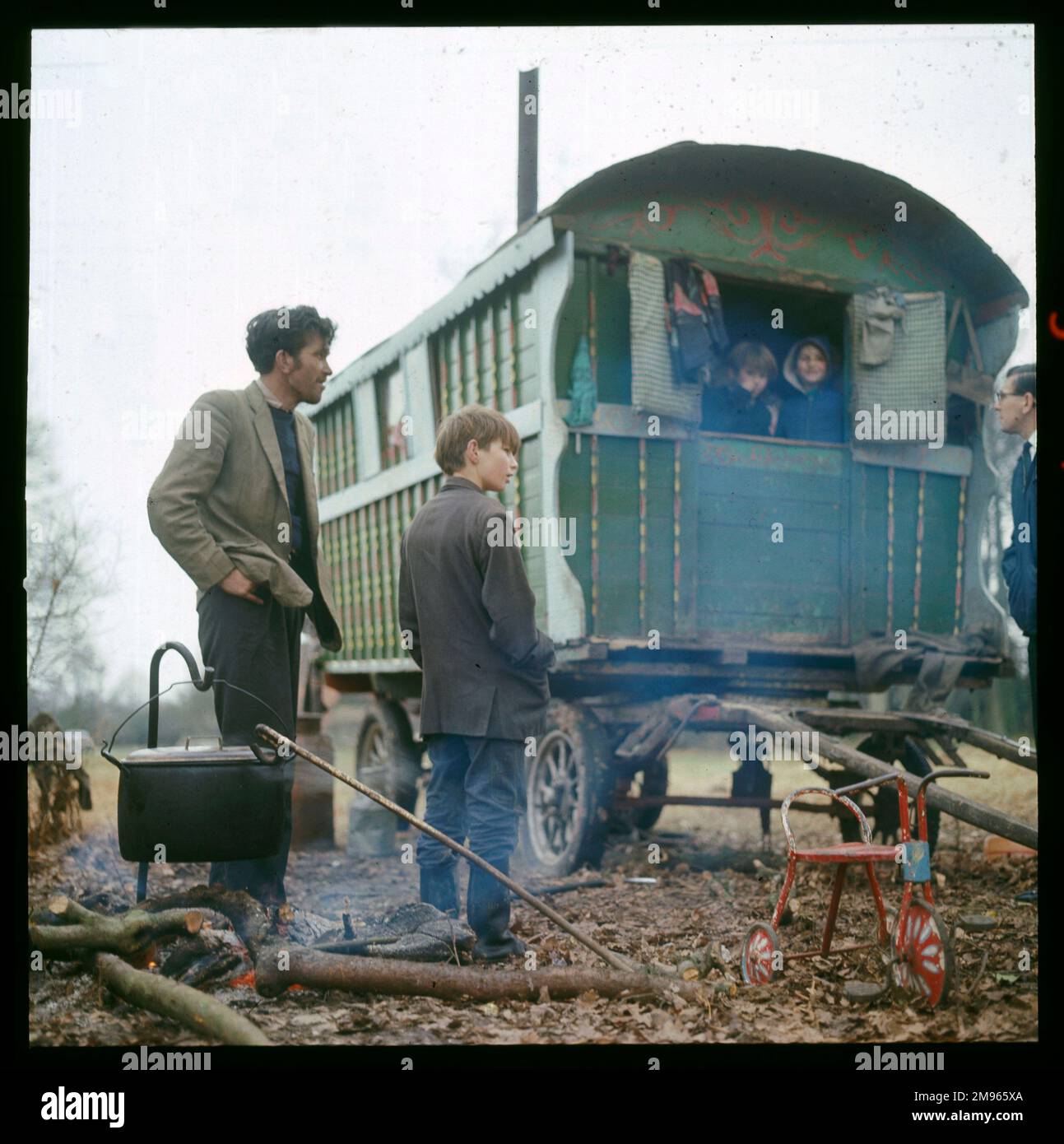 Ein Zigeuner Vater und Sohn stehen neben ihrem grünen Wohnwagen, während ein großer Topf über einem einfachen Feuer kocht. Zwei Kinder schauen aus dem Wohnwagen in diesem Lager in Surrey heraus Stockfoto