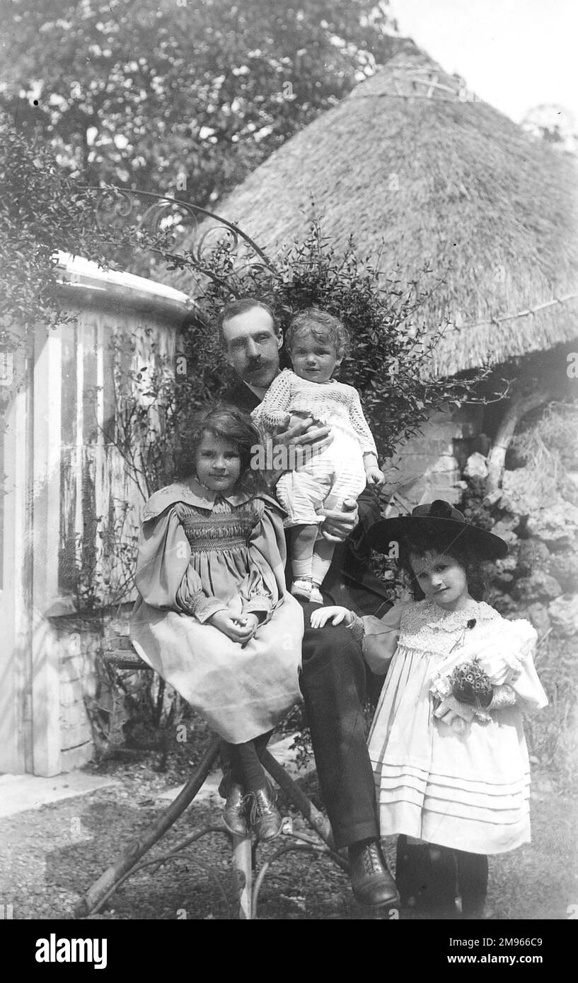 Ein Vater sitzt mit seinen drei Töchtern in einem Garten, eine hält eine Spielzeugpuppe und trägt einen Hut mit breiter Krempe Stockfoto