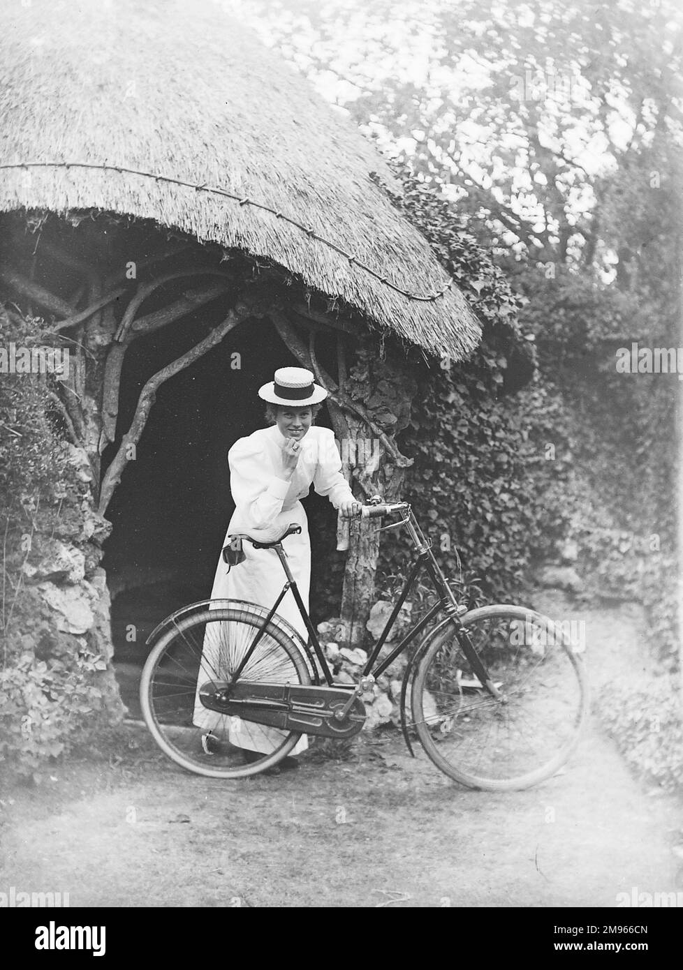 Eine Dame in langem weißem Sommerkleid und Bootsmann lehnt sich auf ihrem aufrechten Fahrrad am Eingang zu einem Sommerhaus oder einer Grotte auf dem Gelände eines großen Landhauses Stockfoto