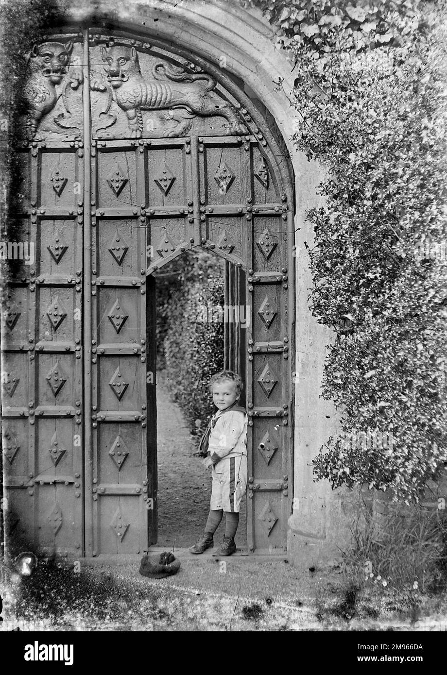Ein kleiner Junge lehnt sich gegen eine Tür, die in eine viel größere Reihe von beeindruckend geschnitzten Holztoren geschnitten wurde. Er trägt einen Seemannsanzug, hat aber seine Mütze weggeworfen (Abbildung 2 von 3) Stockfoto