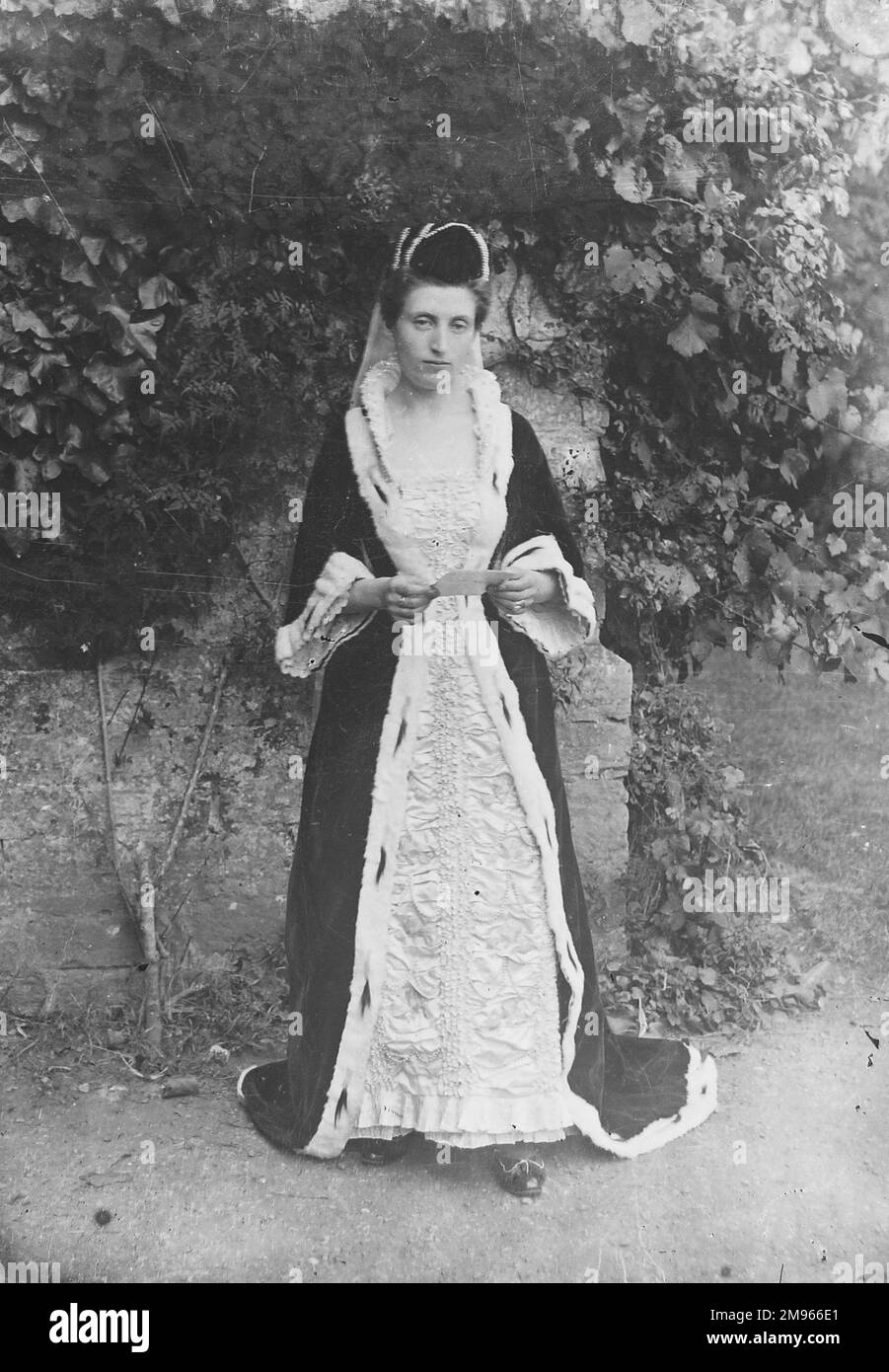 Eine Dame in mittelalterlichen Kostümen mit fließenden Bademänteln und Schleier für eine Party oder eine Vorstellung. Sie posiert an der großen Wand eines großen Gartens unter dem Efeu und hält eine Karte Stockfoto