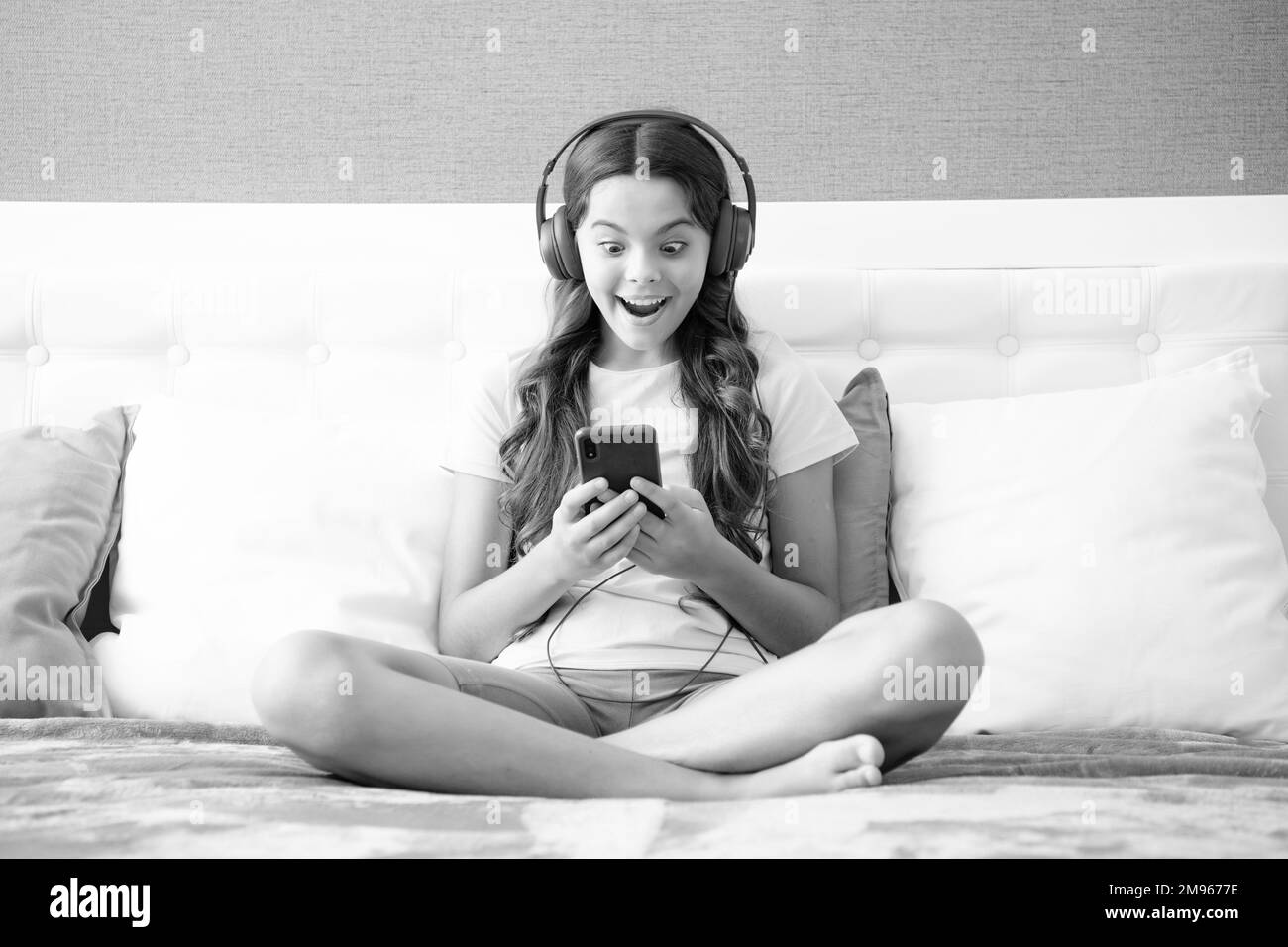 Überraschendes Gesicht, Überraschungsemotionen von Teenager-Mädchen. Teenager Kind Mädchen tragen Kopfhörer Videos hören Musik auf Smartphone sitzen auf Stockfoto