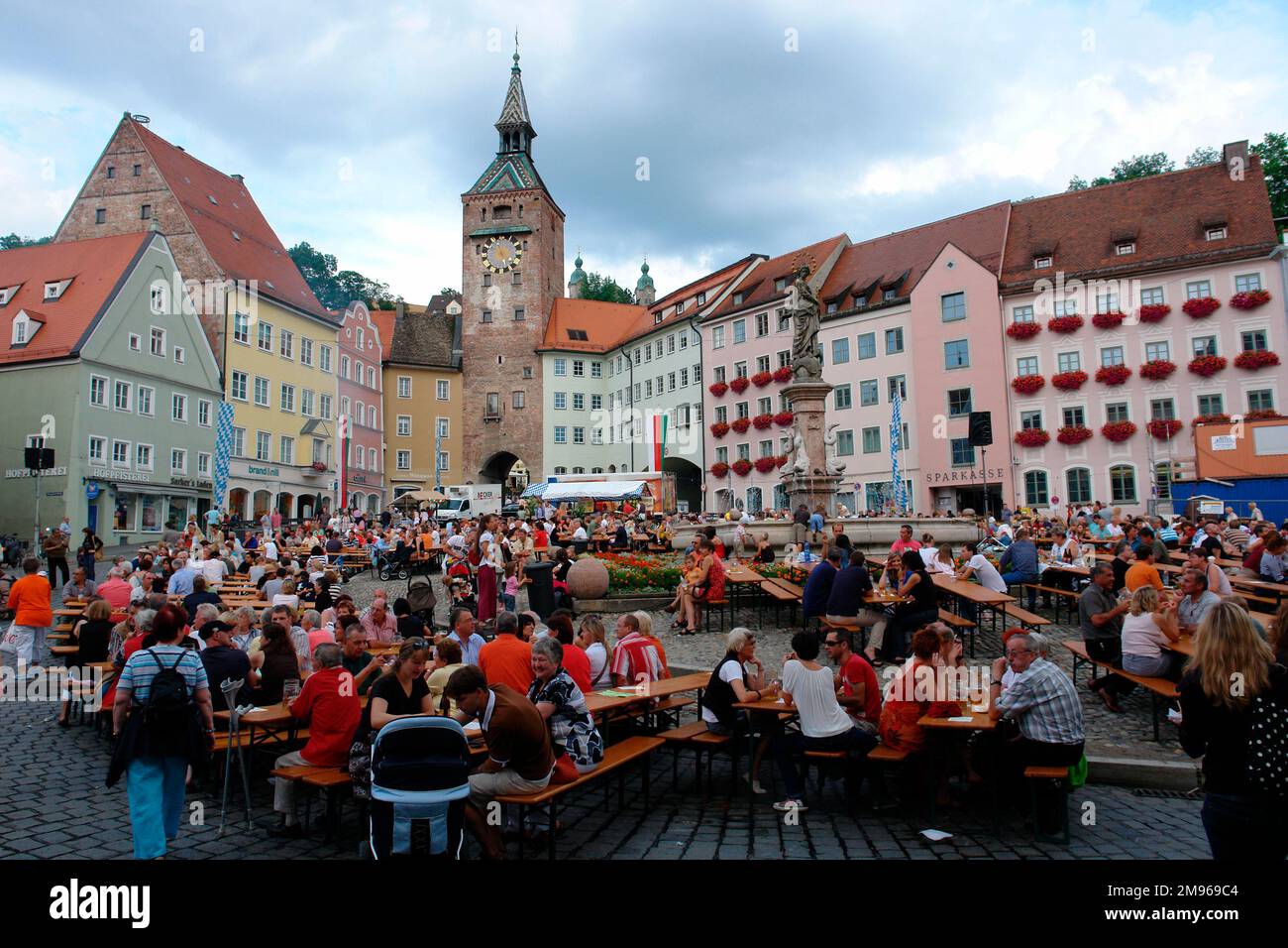 Wochenendfestival auf dem malerischen Marktplatz Landsberg am Lech, Bayern, mit Menschen, die an Böschungstischen sitzen. Der Schönestürm (wunderschöner Turm) neben dem alten Rathaus ist links vom Zentrum im Hintergrund zu sehen. Stockfoto