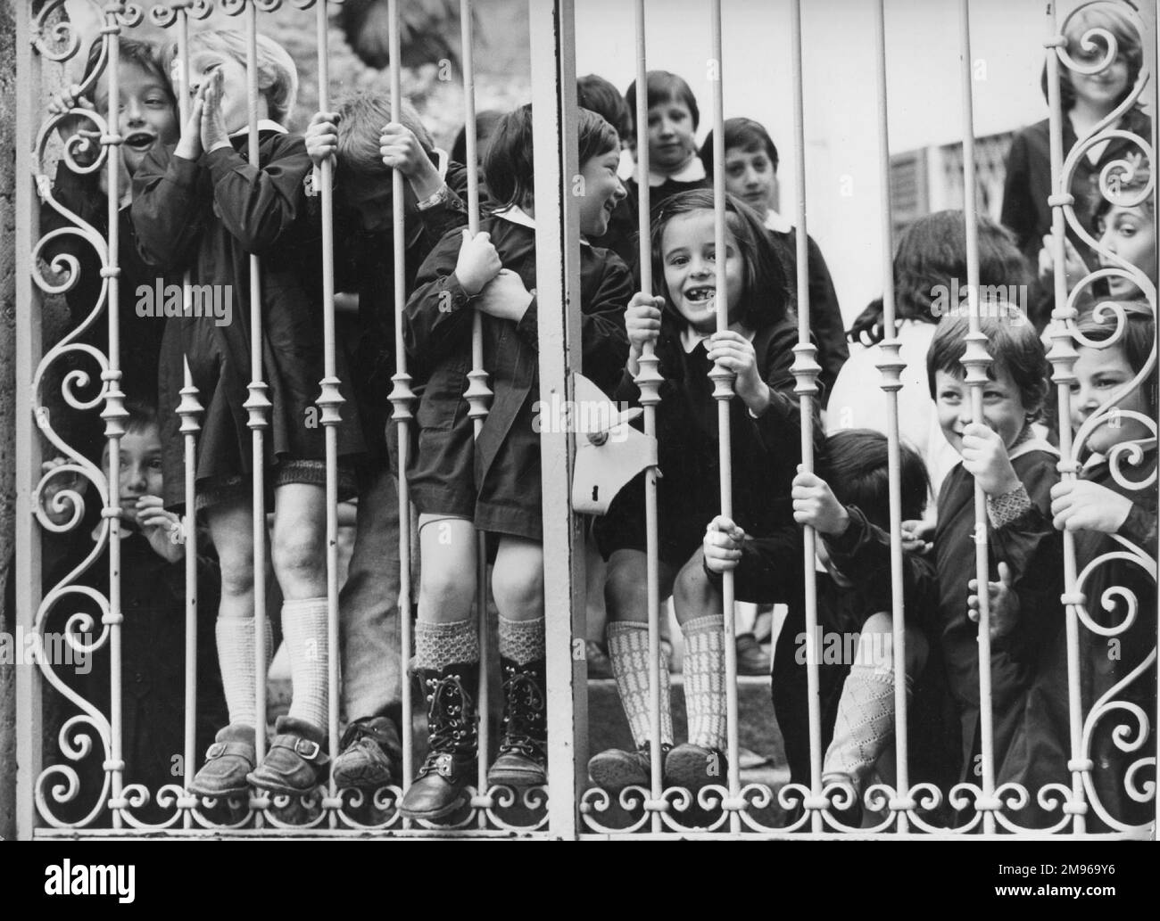 Eine glückliche Gruppe von Kindern, die durch ihre Schultore schauen, wahrscheinlich aufgeregt, fotografiert zu werden. Ihr Lehrer lächelt im Hintergrund rechts. Stockfoto