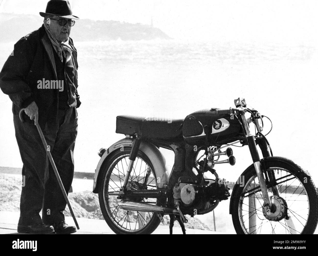Ein alter Mann mit einem Gehstock, der neben einem Motorrad steht, das in der Nähe eines Strandes geparkt ist. Stockfoto