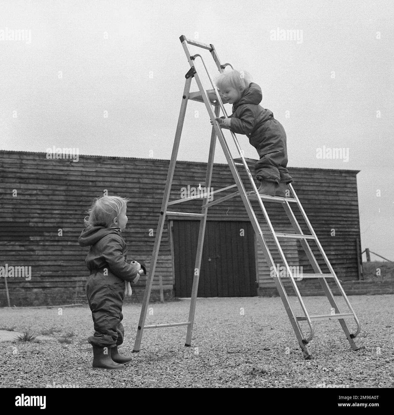 Zwei Kleinkinder, die mit einer Stehleiter auf einer Kiesfläche vor einem Bauernhaus spielen. Stockfoto