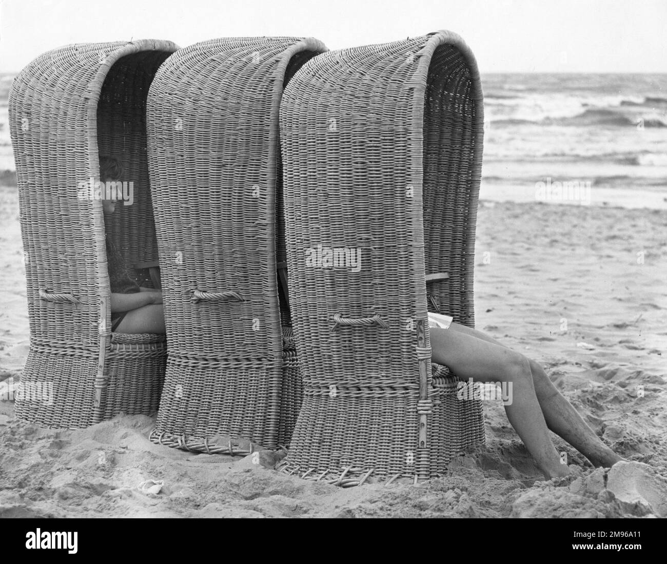 Drei große Basketballliegen an einem belgischen Strand, in denen zwei Personen sitzen. Sie wurden entwickelt, um maximalen Schatten vor der heißen Sonne zu bieten. Stockfoto