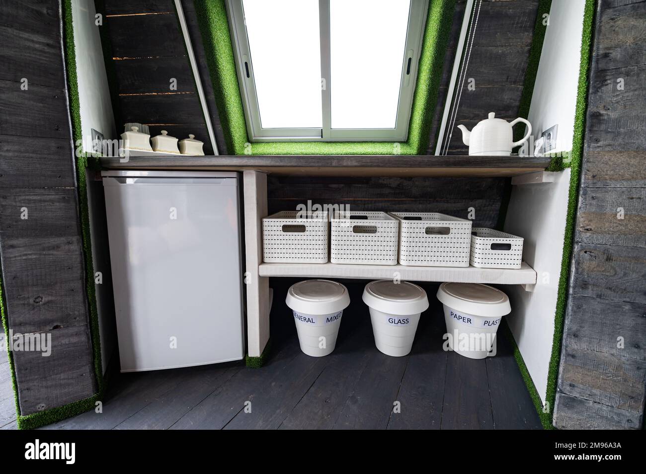 Kleine Küchenelemente in einem Holzsommerhaus. Kühlschrank, Wasserkocher, heiße Getränke, Geschirr und Recyclingdosen in einer kleinen Küche. Originell, ungewöhnlich, vielleicht Stockfoto