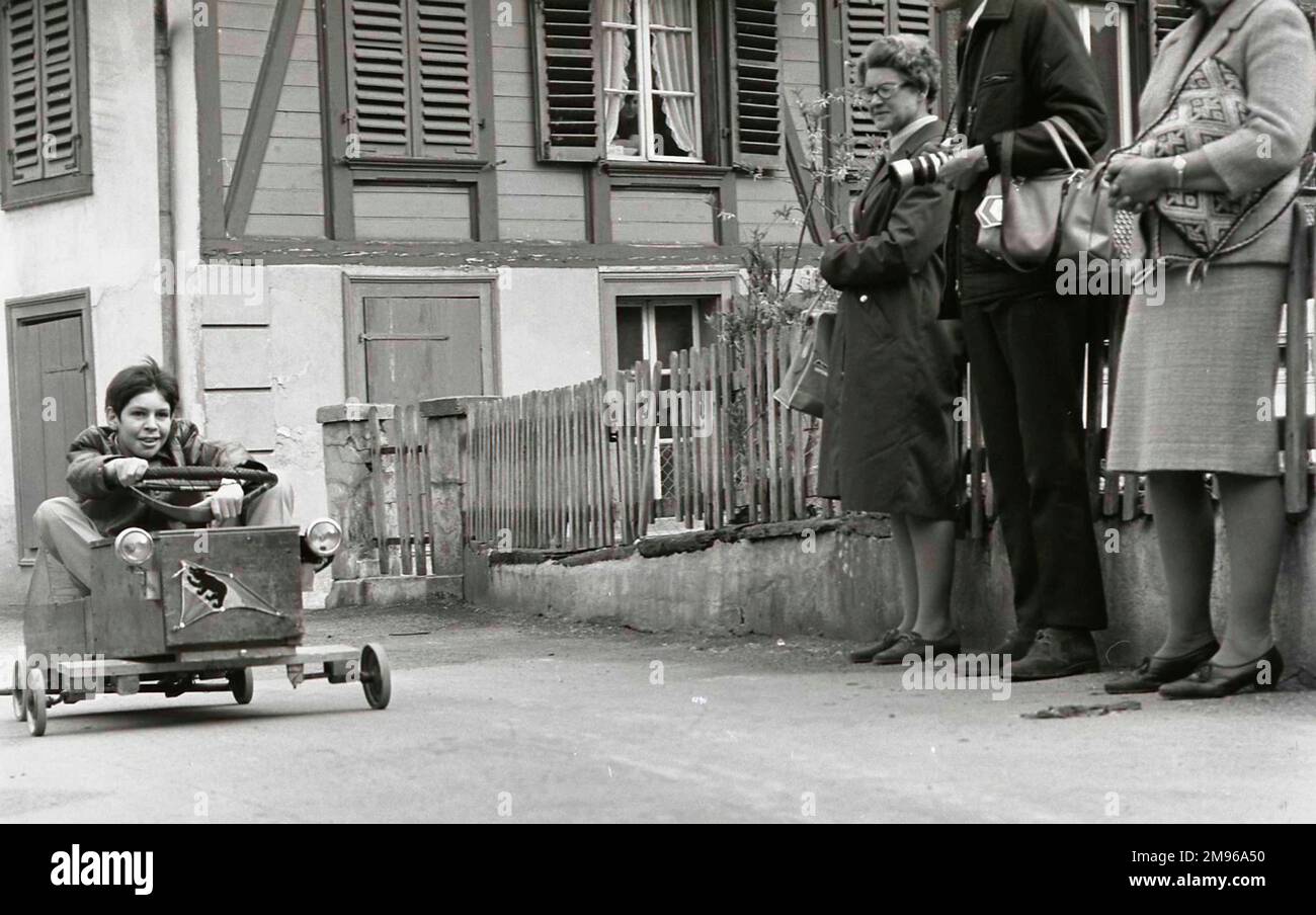 Ein Junge fährt mit seinem Gokart, beobachtet von drei Erwachsenen, die aussehen, als würden sie auf einen Bus warten. Stockfoto