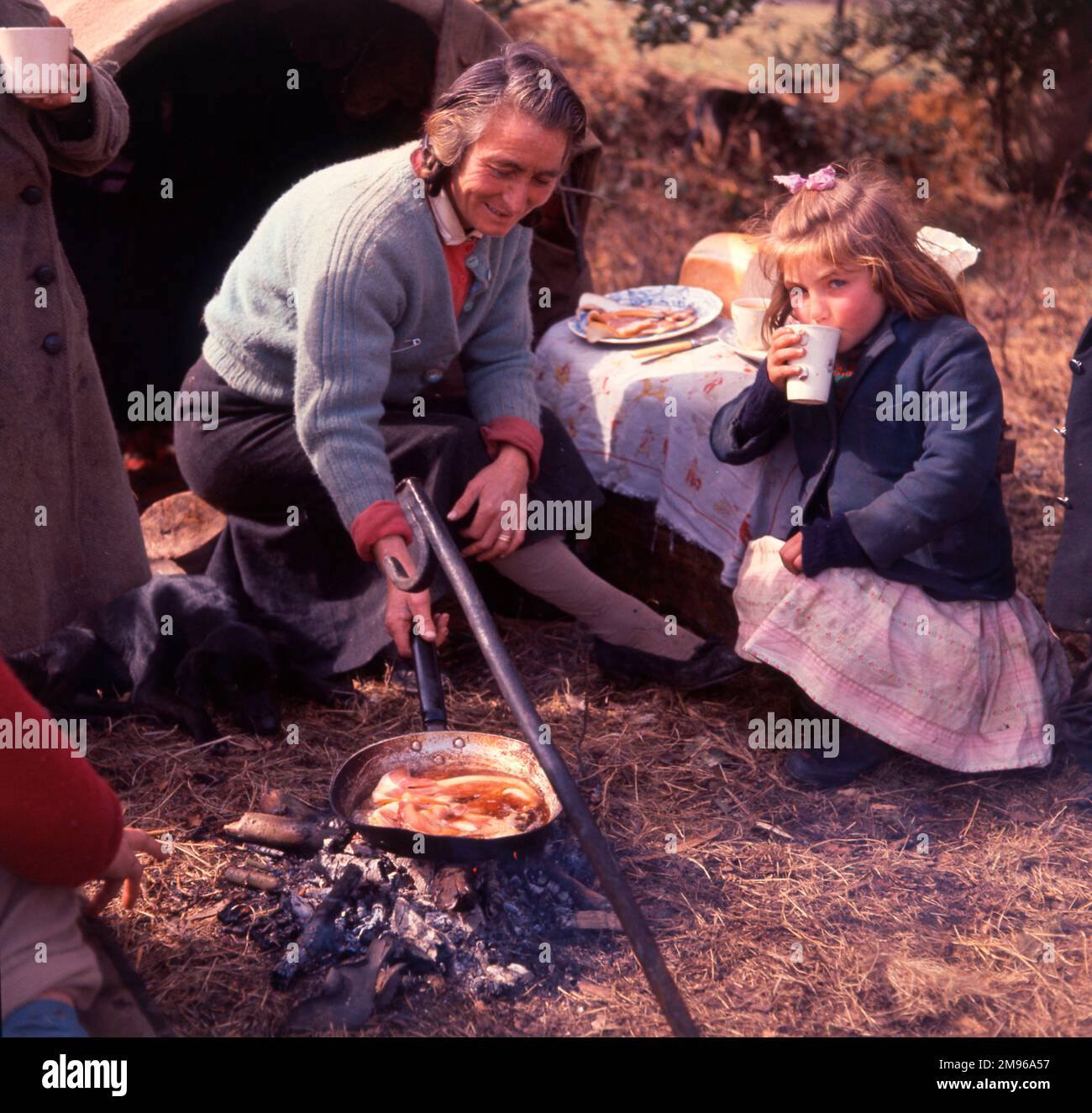 Eine Zigeunerin, die im Freien ein gebratenes Frühstück kocht. Ein kleines Mädchen hockt in der Nähe und schlürft eine Tasse Tee. Stockfoto