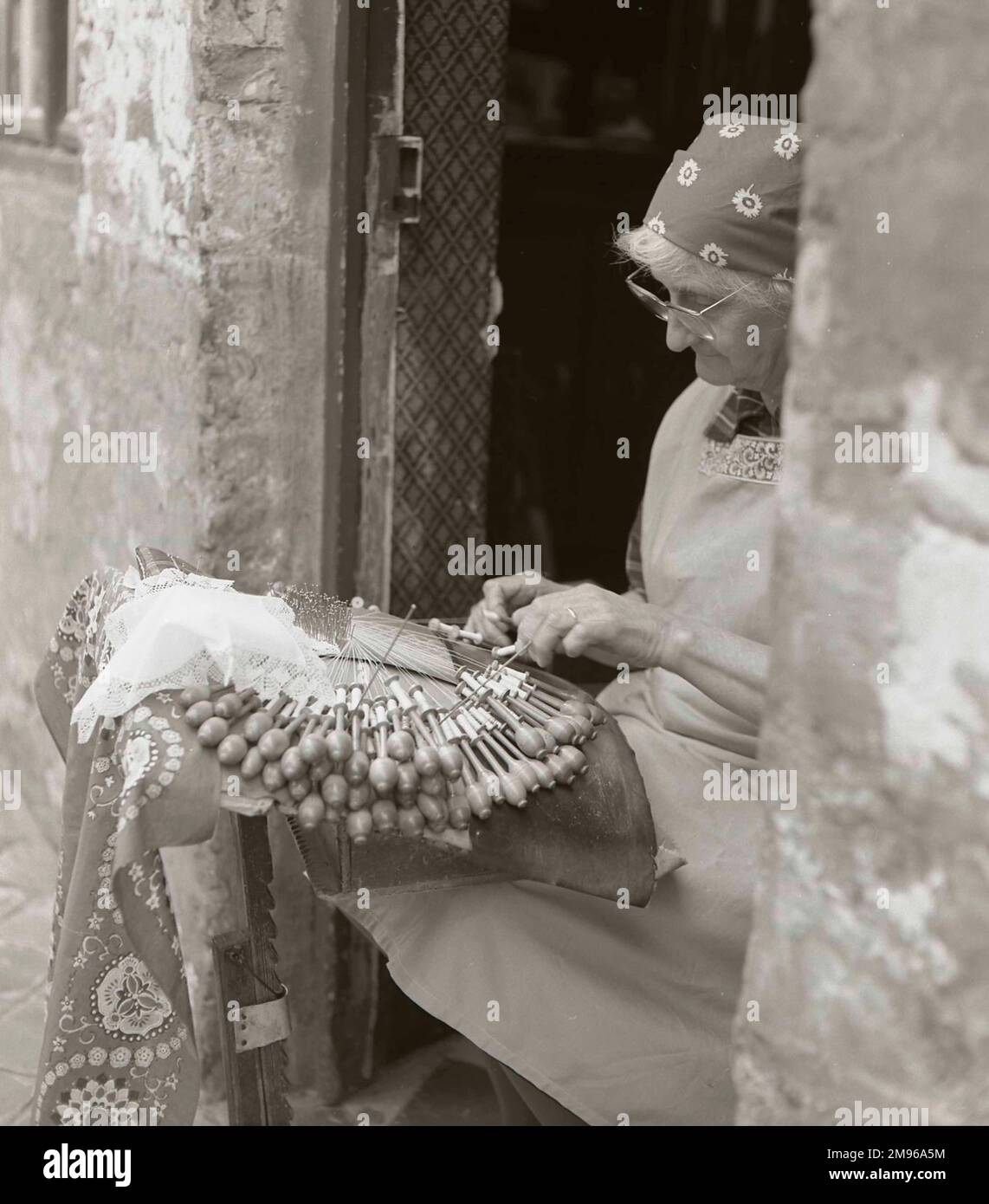 Eine ältere Frau, die in der offenen Tür ihres Hauses Spitzen herstellt, mit einer großen Anzahl von Spulen. Stockfoto