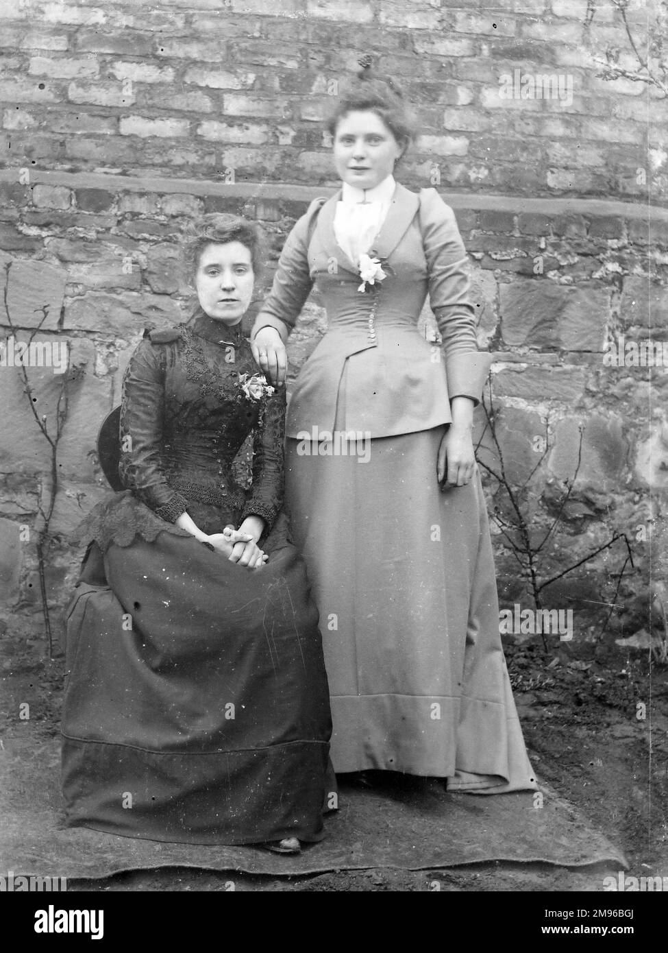 Zwei Schwestern posieren für ihr Foto vor einer Gartenmauer, wahrscheinlich in der Gegend von Mid Wales. Sie tragen lange Röcke und eng geschnittene Jacken. Stockfoto