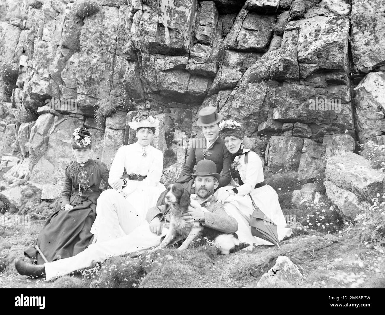 Eine edwardianische Gruppe am Meer (vier Frauen und ein Mann), die vor einigen Felsen saß. Der Mann hält einen Hund in der Hand, der eifrig wegzurennen scheint. Stockfoto
