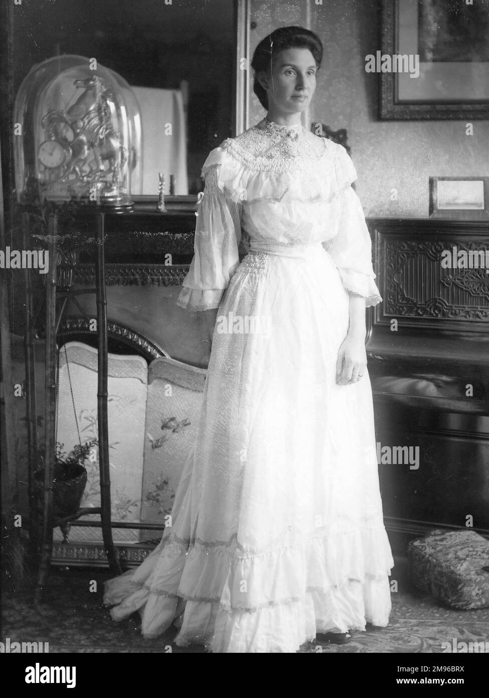 Eine elegante Dame in einem durchgehenden weißen Kleid posiert in einem Landhaus in Mid Wales. Es gibt einen Kamin und dekorierten Bildschirm auf der linken Seite und ein aufrechtes Klavier auf der rechten Seite. Stockfoto