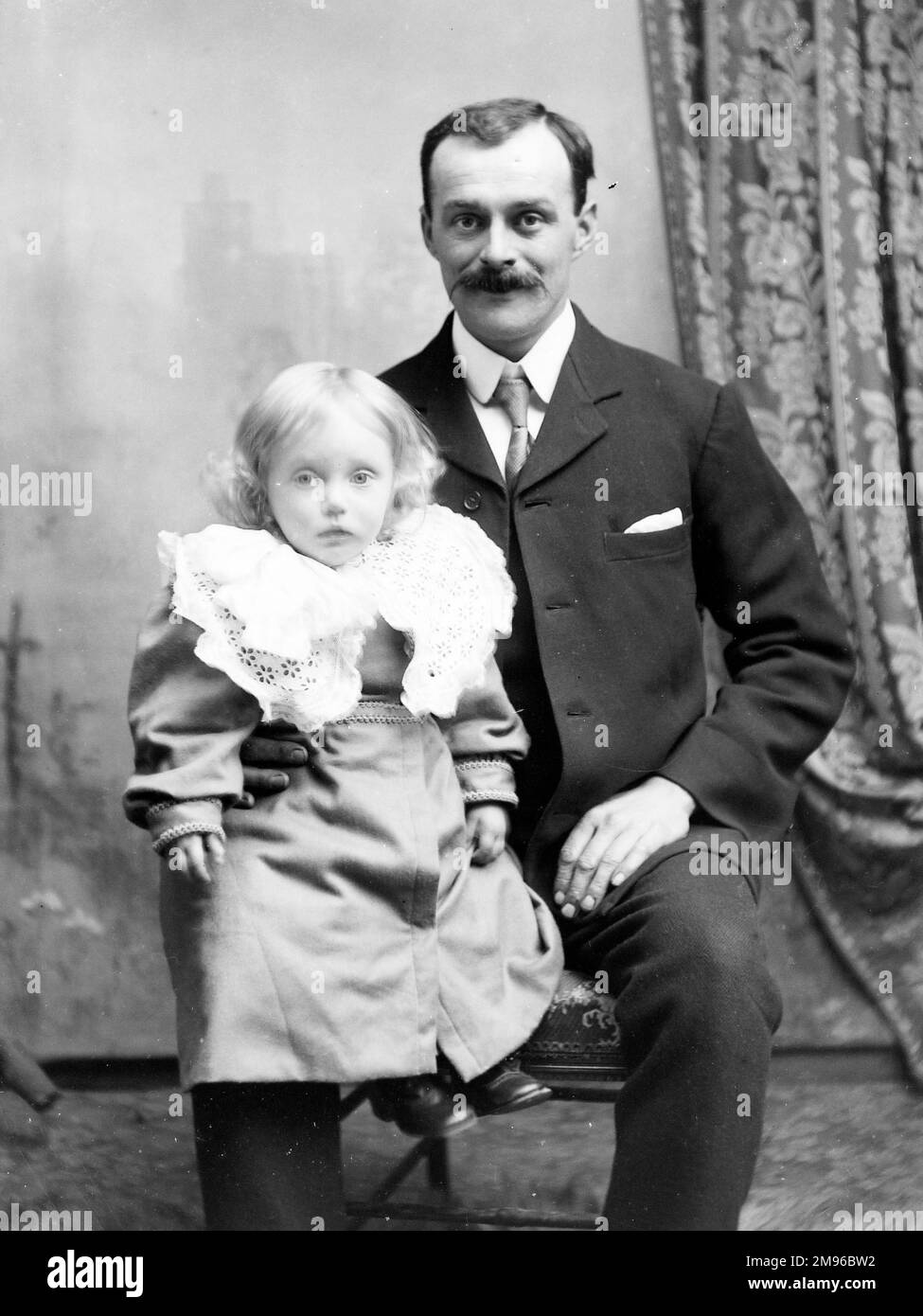 Ein Vater mit seiner kleinen Tochter, die im Studio des Fotografen, Mid Wales, auf dem Knie saß. Es gibt eine bemalte Kulisse mit einem Vorhang an der Seite. Stockfoto