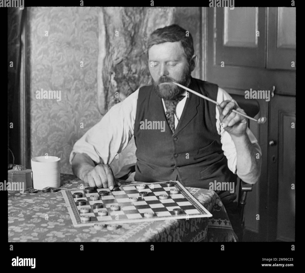 Ein Mann rätselt über ein Spiel von Draghts, das er anscheinend alleine spielt. Er hält eine lange Kirchenpfeife in seinem Mund. Stockfoto Ein Mann rätselt über ein Spiel von Draghts, das er anscheinend alleine spielt. Er hält eine lange Kirchenpfeife in seinem Mund. Stockfoto