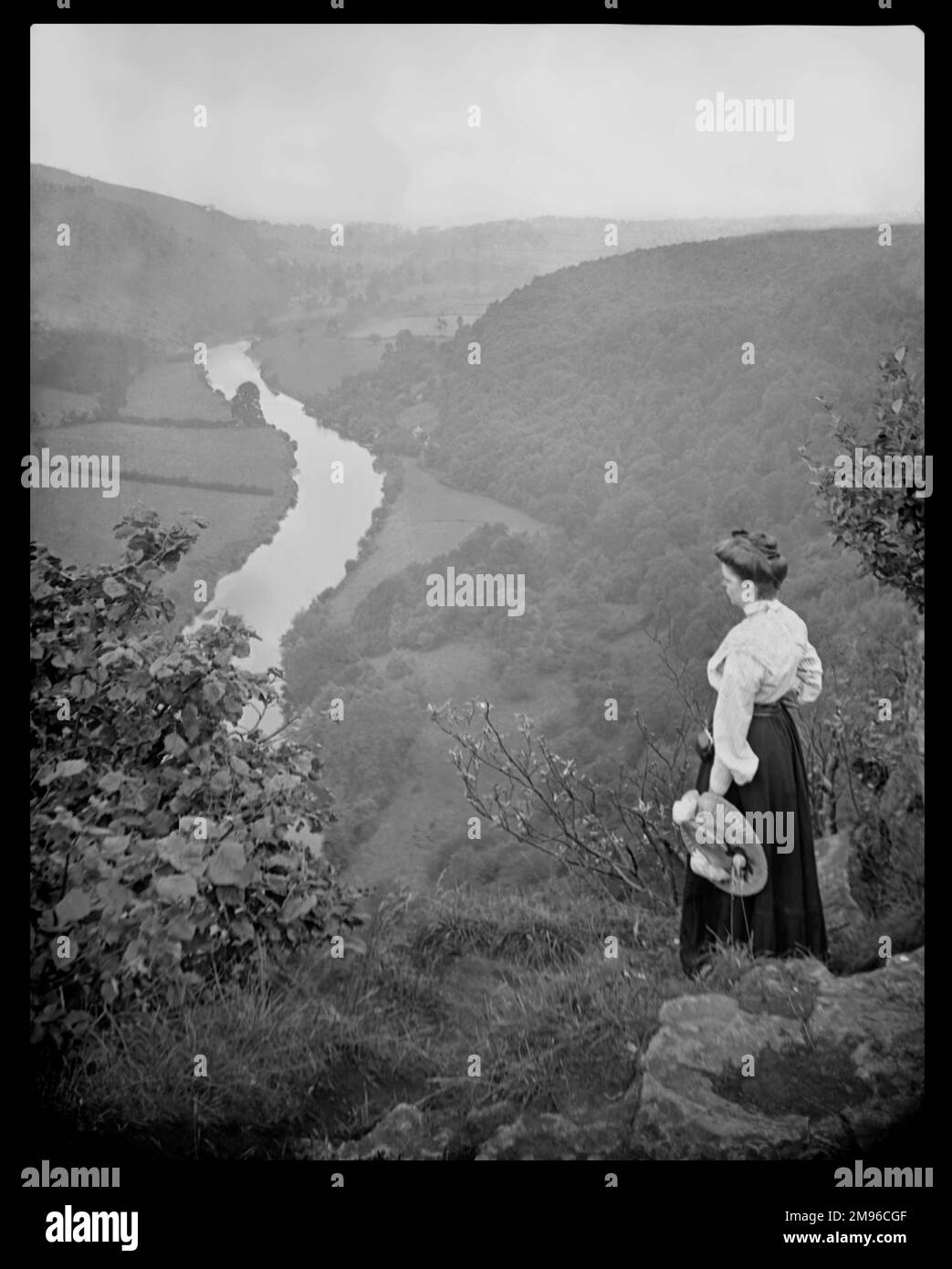 Eine edwardianische Frau, die in ein Tal zwischen sanften Hügeln schaut, wo ein Bach fließt. Stockfoto